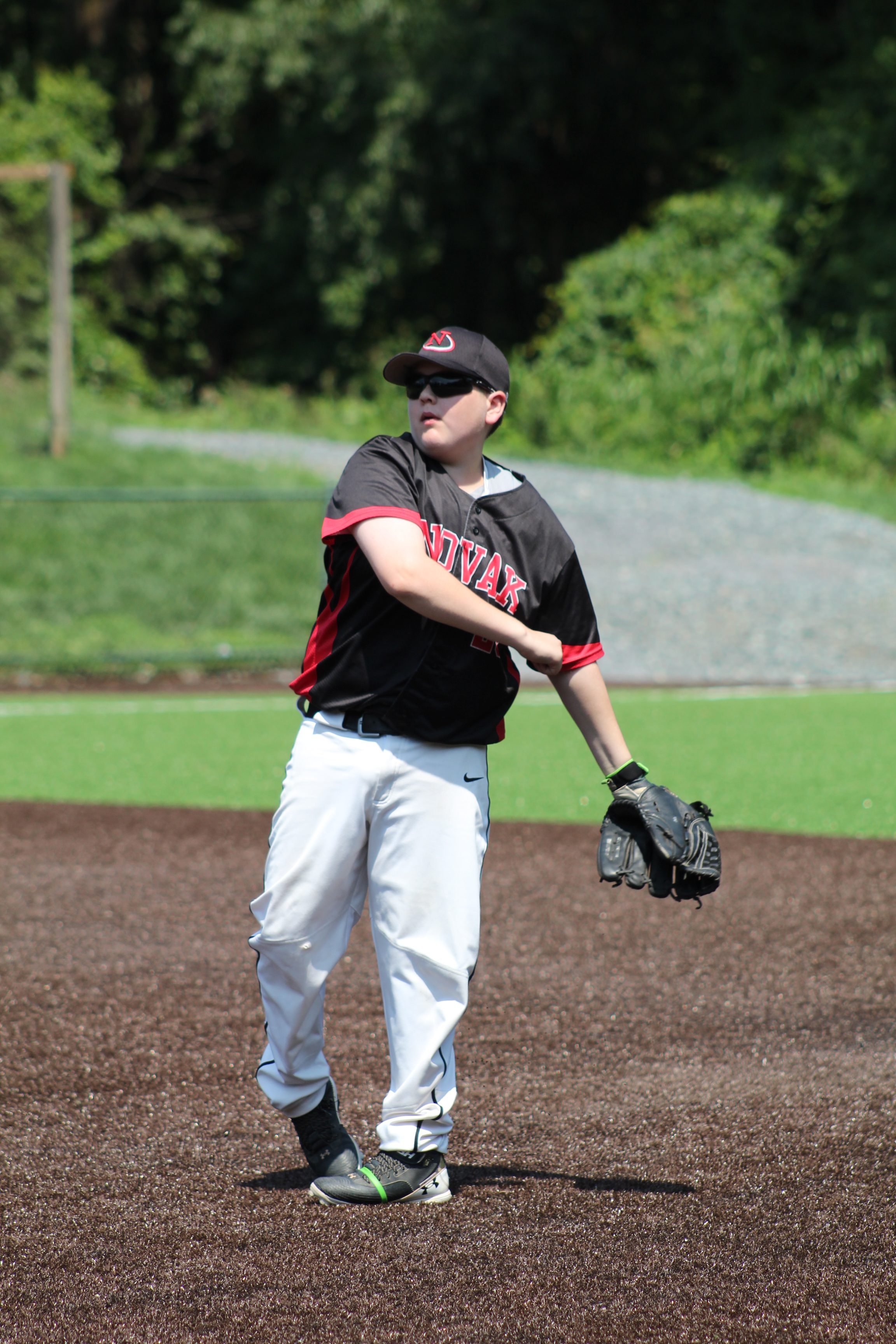 A young baseball player in a black jersey and white pants prepares to throw a baseball while wearing sunglasses and a cap.