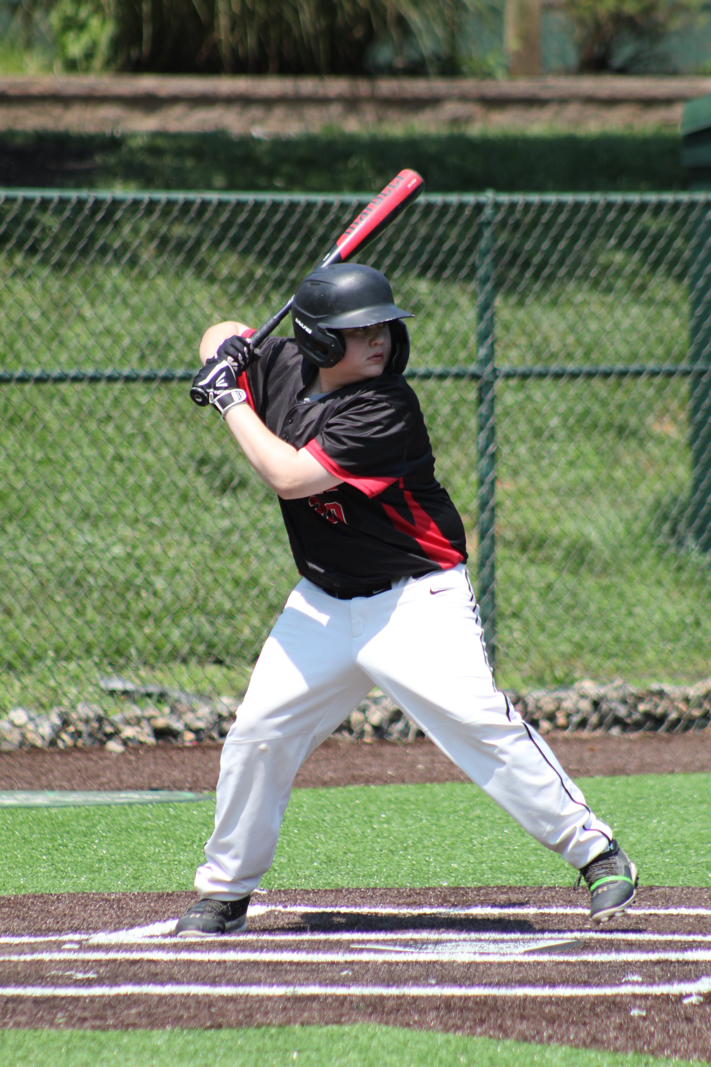 A young baseball player in a black jersey and white pants stands in a batting stance, holding a red bat, on a grassy field with a chain-link fence in the background.