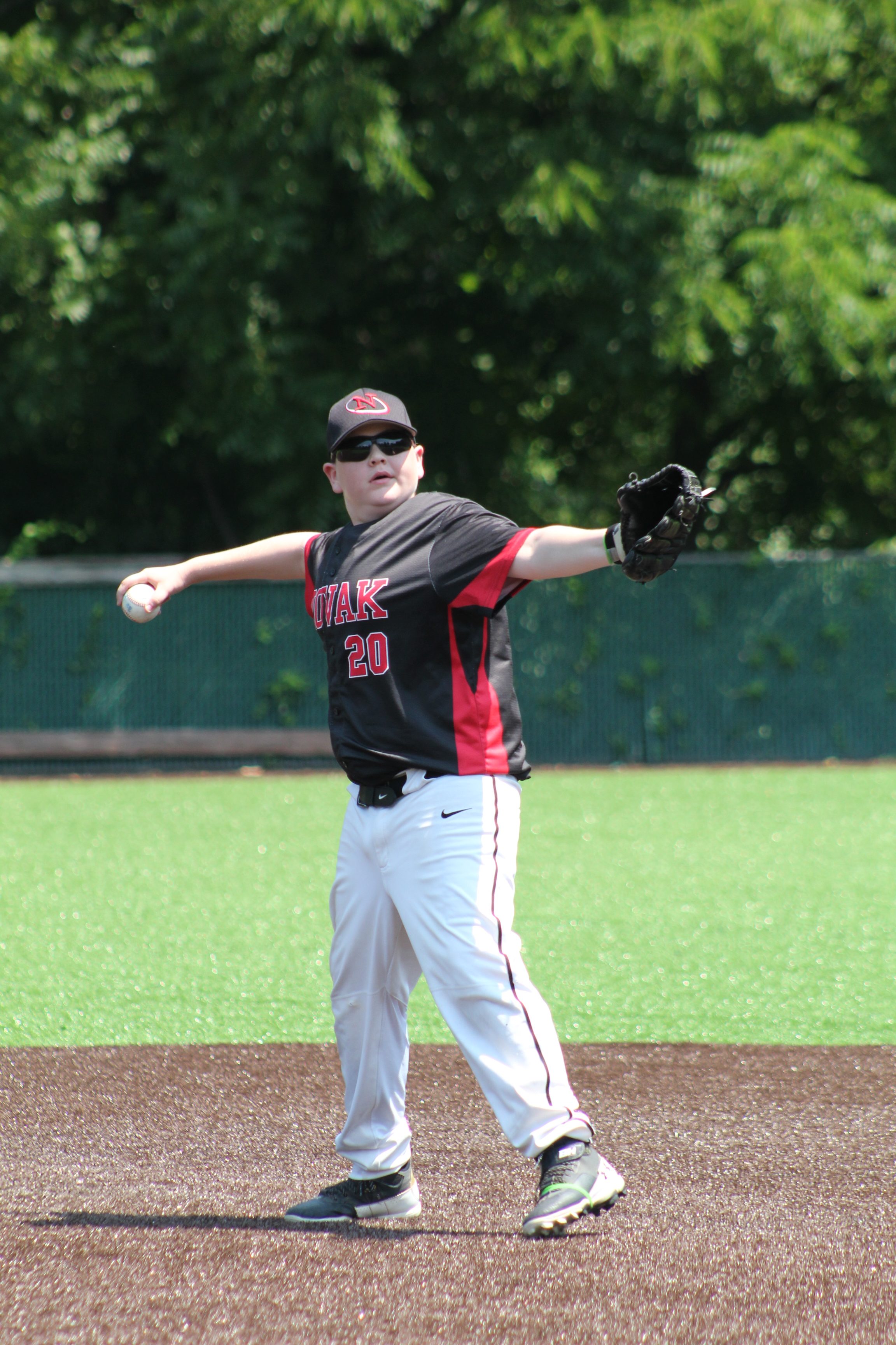 A young baseball player wearing a black and red jersey and white pants is preparing to throw a baseball while standing on a field with artificial turf.
