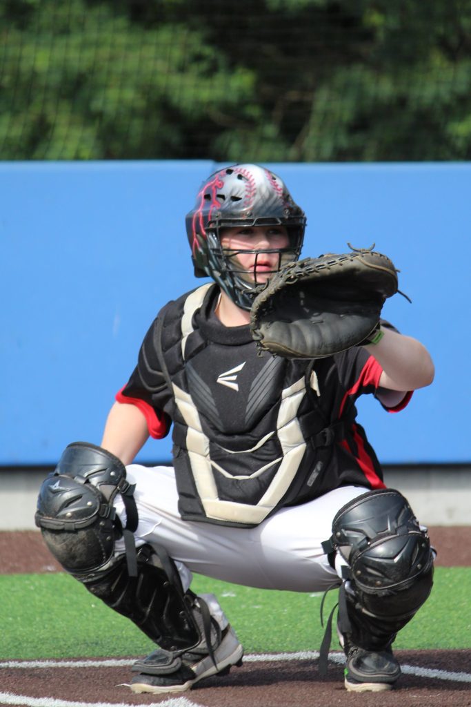 A youth baseball catcher in full gear, wearing a helmet and chest protector, crouching and signaling on the field.