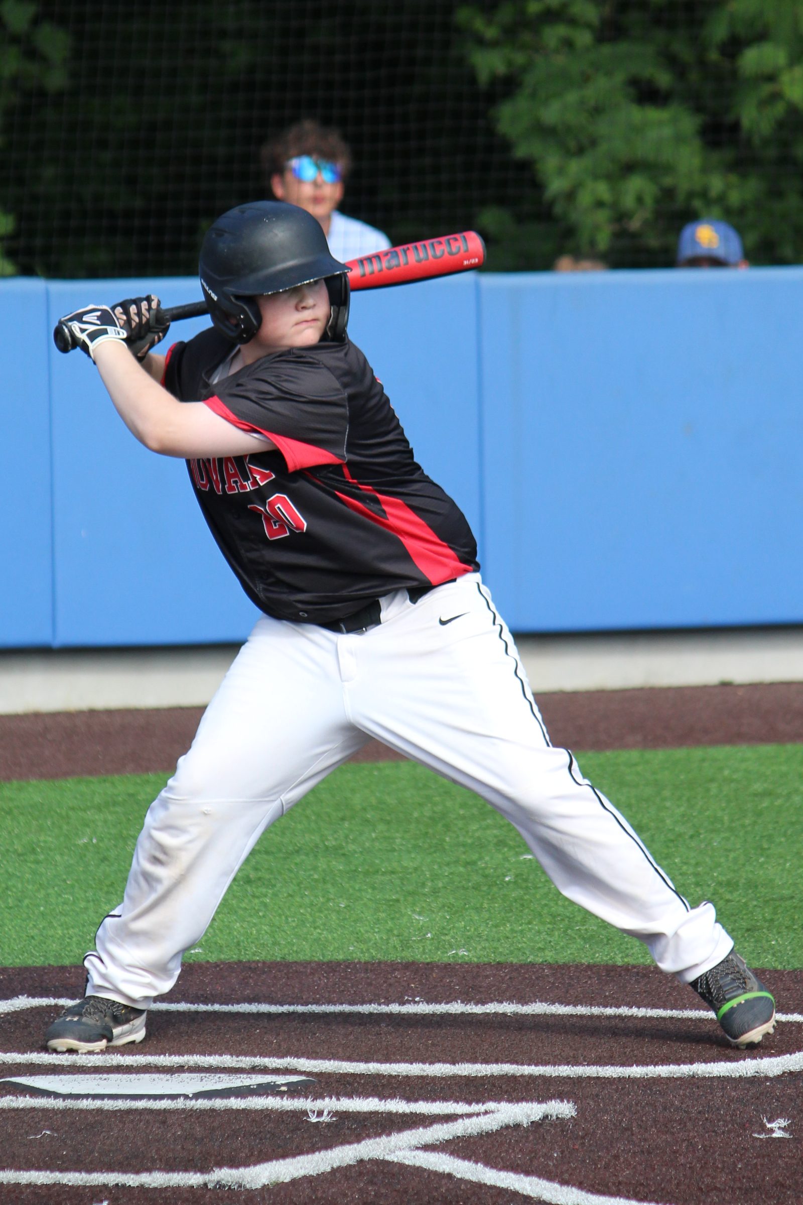 A young baseball player in a black and red uniform stands poised with a bat, ready to swing. The player wears a helmet and is on a baseball field with a blue background and spectators in the distance.
