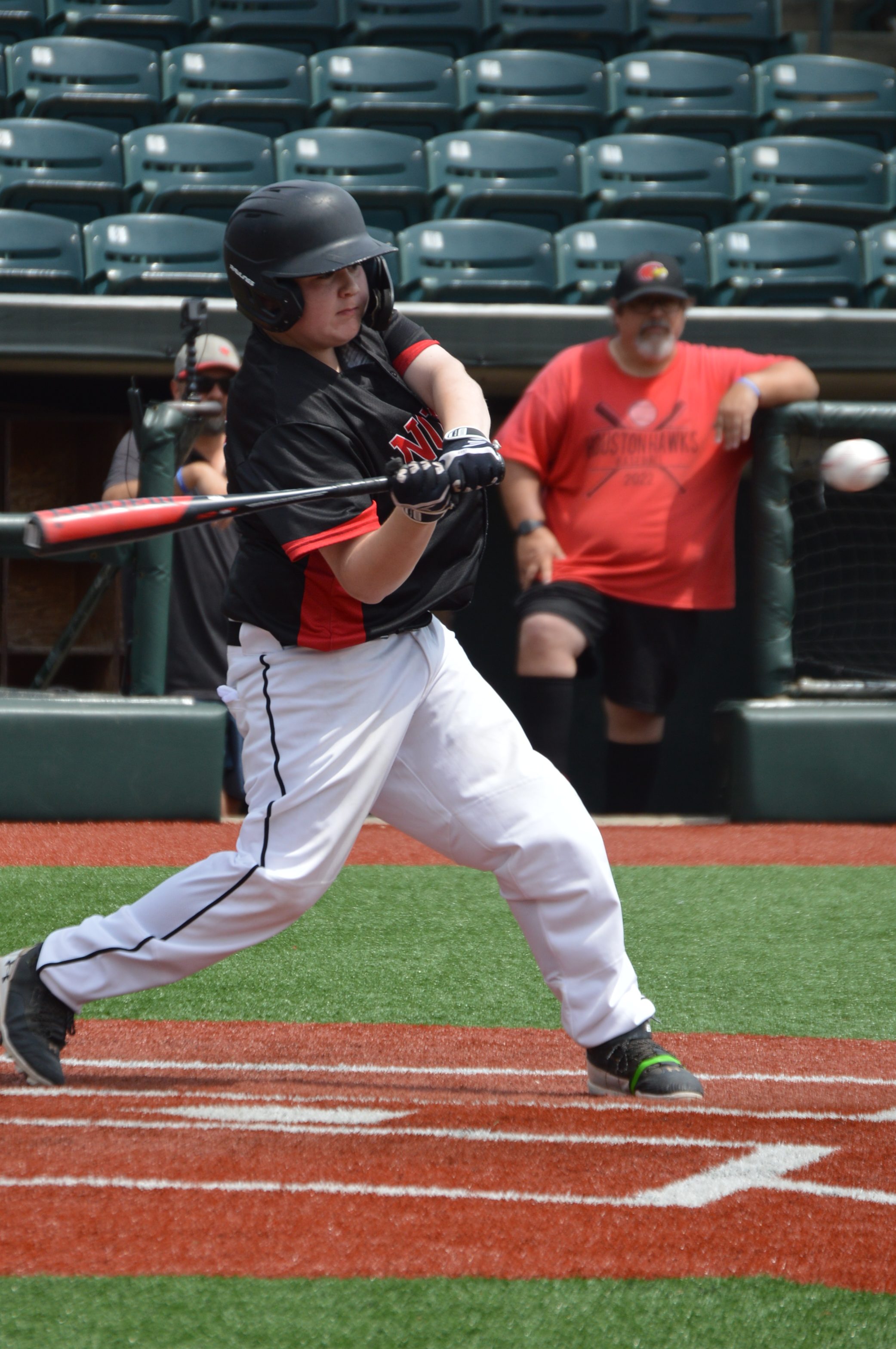 A young baseball player in a black and red uniform swings a bat at home plate, with a blurred baseball in motion. Spectators can be seen in the background.
