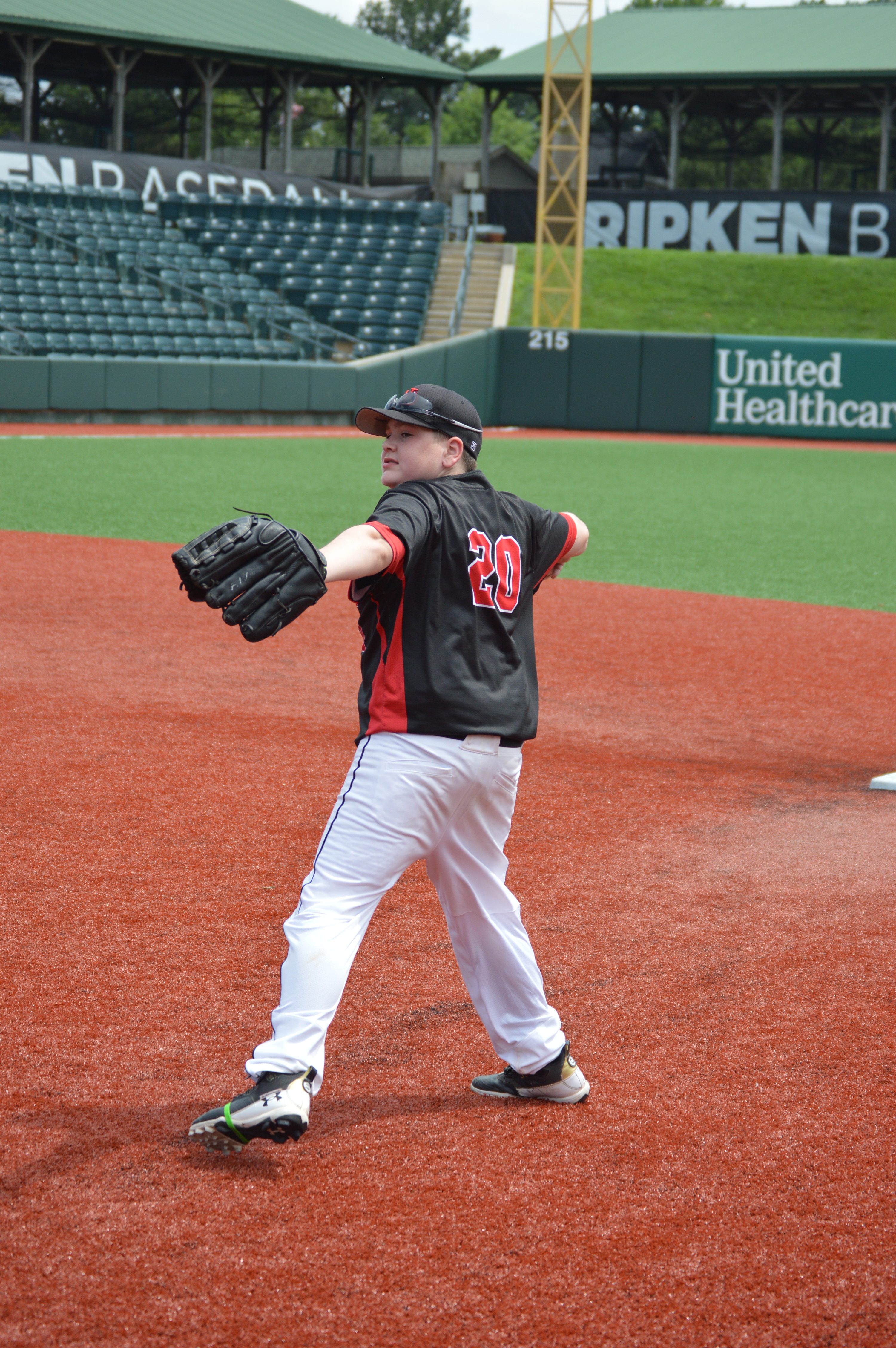 A young baseball player in a black and red uniform is preparing to throw a baseball on a field with a turf surface, with empty stadium seating in the background.
