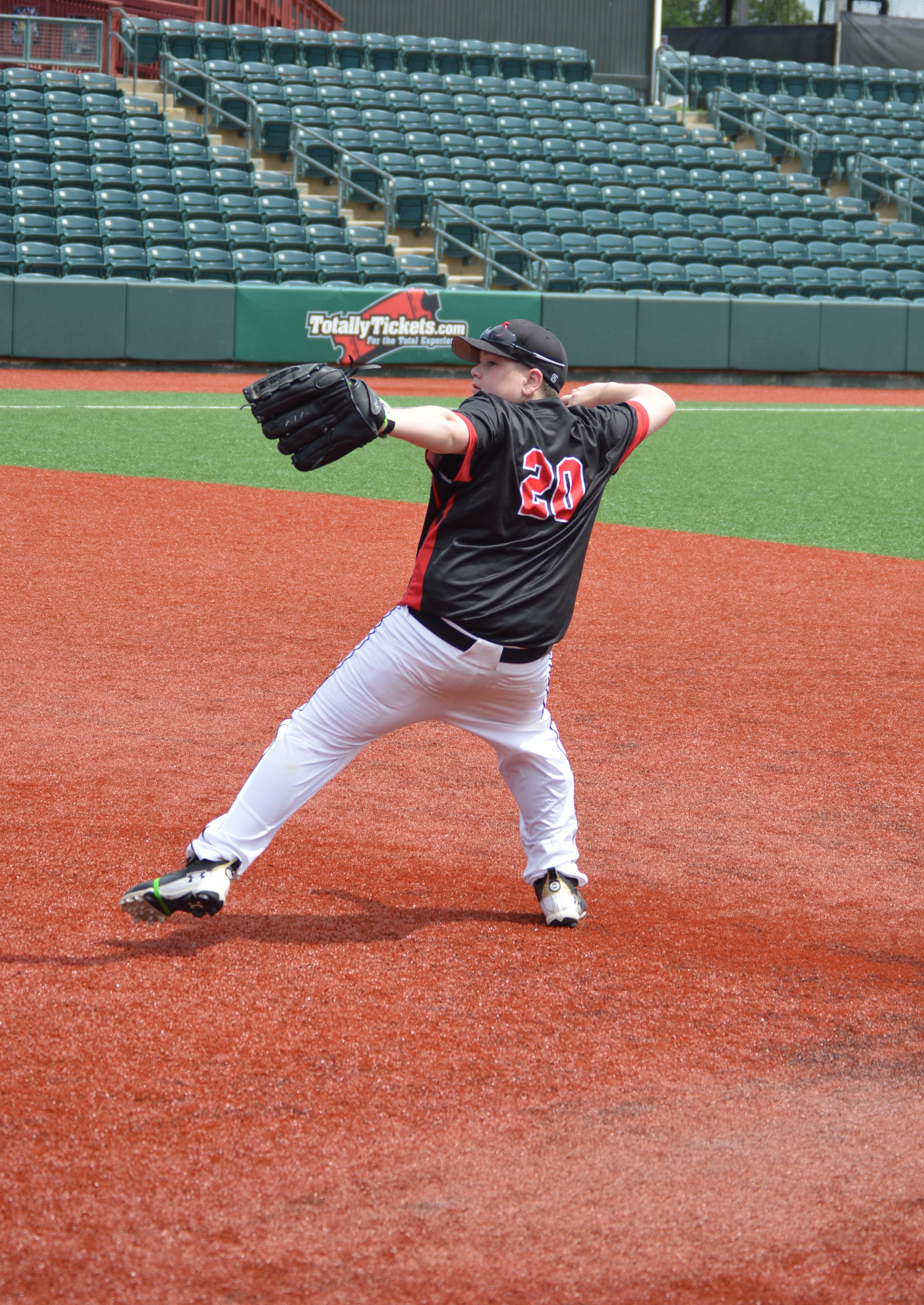 A young baseball player in a black jersey with the number 20 is in the process of pitching on a baseball field with a red turf surface and empty green seating in the background.