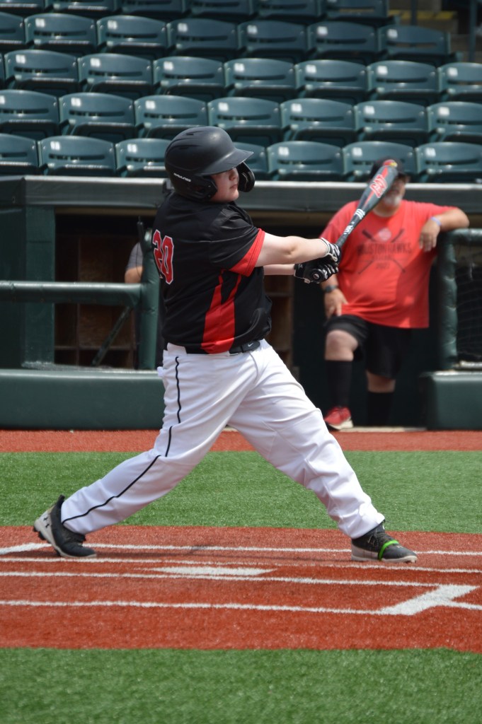 A young baseball player in a black and red uniform swings a bat on the field, with a blurred audience in the background.