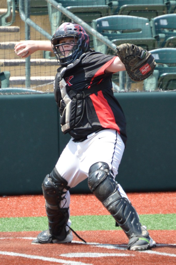 A young baseball catcher in a black and red uniform prepares to throw the ball while on the field.