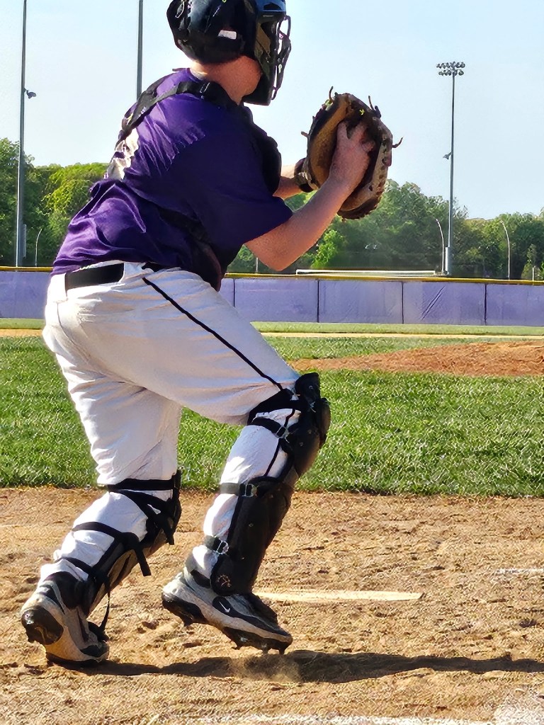 A catcher in a purple jersey prepares to receive a pitch on a baseball field, wearing protective gear and readying his glove.