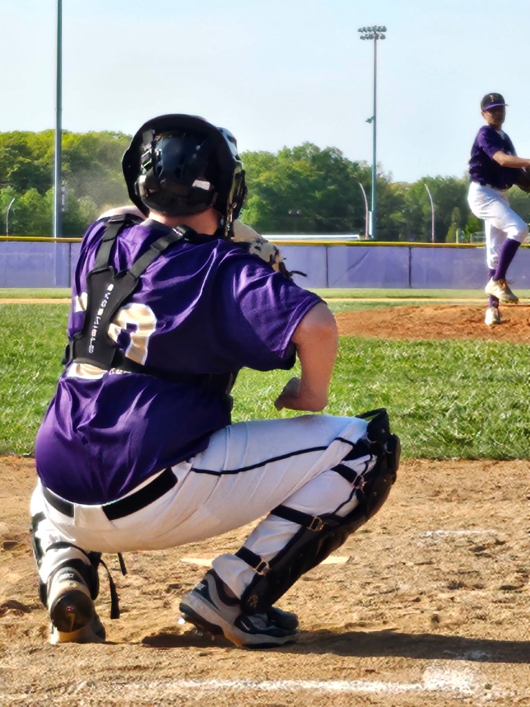 A baseball catcher in a purple jersey squatting behind home plate, watching a pitcher prepare to throw.