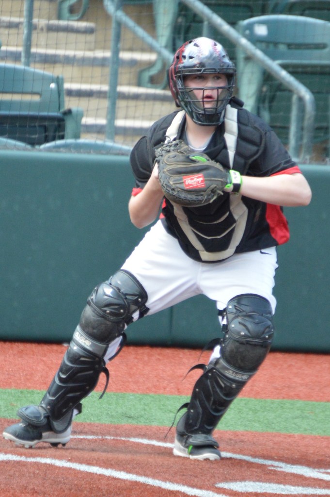 A young baseball catcher in full gear, including a helmet and chest protector, is poised in a catching stance on a baseball field.