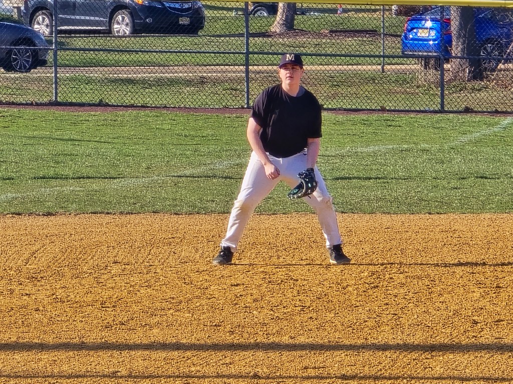 A young player in a black shirt and baseball cap stands in a ready position on a baseball field, with a dirt infield and green grass in the background.