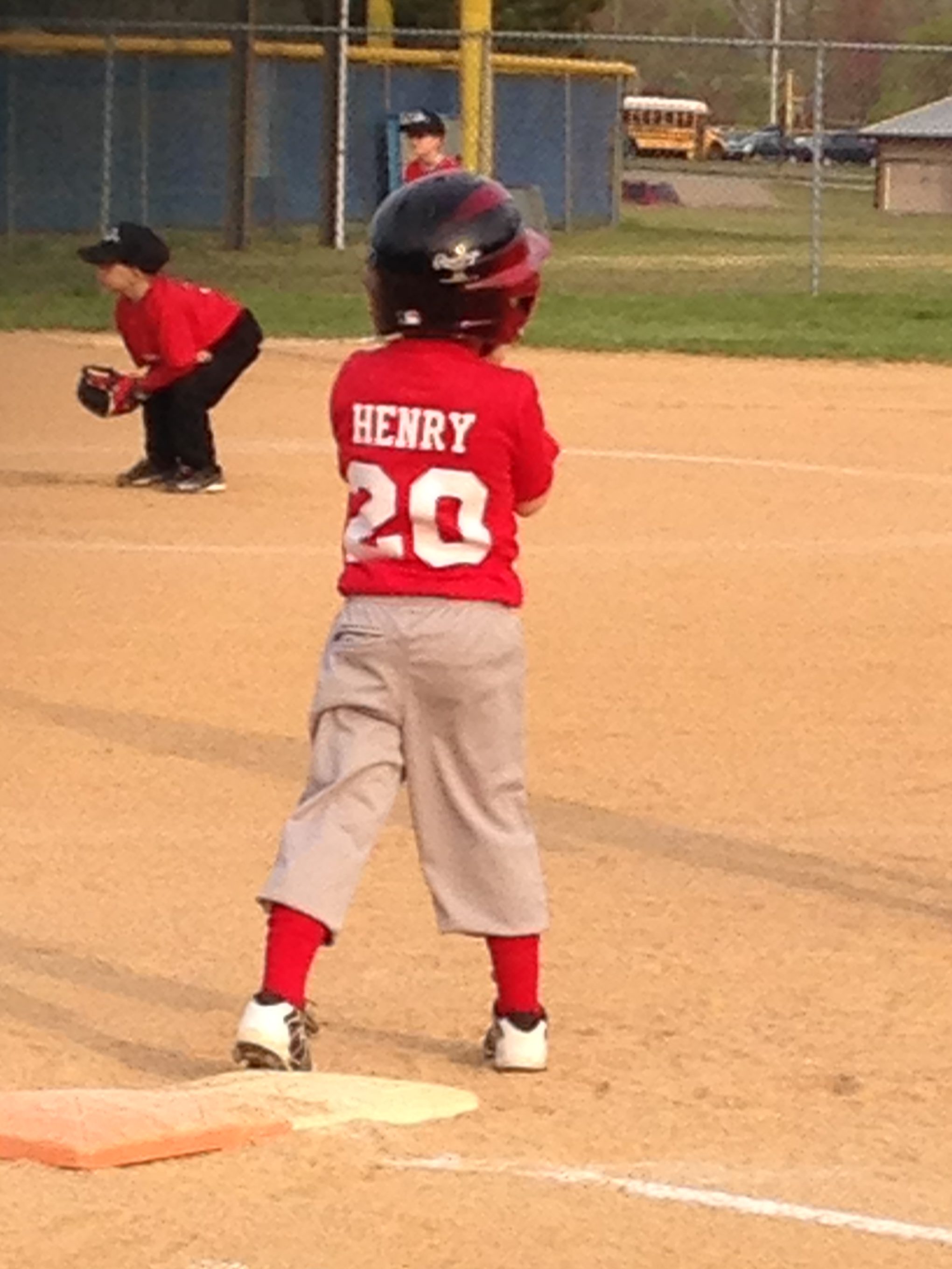 A young baseball player wearing a red jersey with the name 'HENRY' and the number '20' stands on the field, preparing to bat, with a baseball cap and helmet on. In the background, another player crouches in a ready position.
