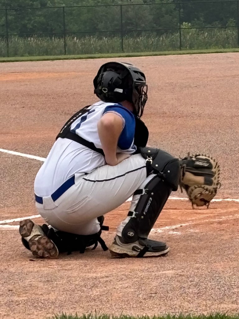 A baseball catcher crouching on the field, wearing protective gear and a jersey, ready for play.