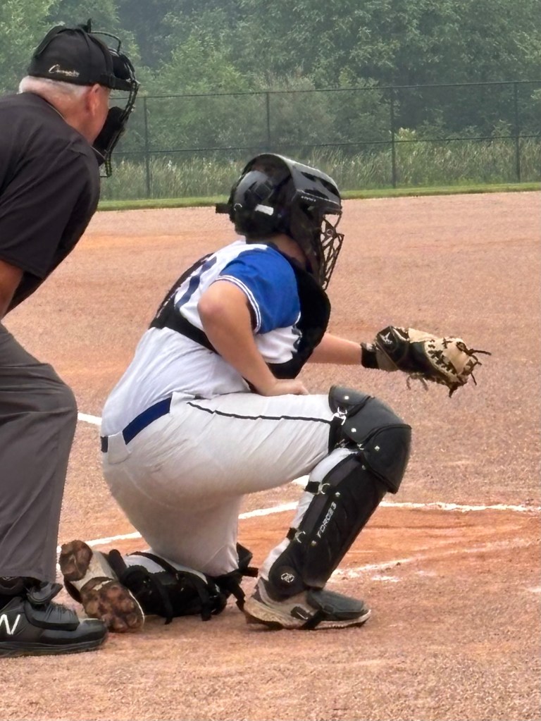 A youth baseball catcher wearing a helmet and chest protector crouches behind home plate, preparing for the pitch, while an umpire stands in front of him.