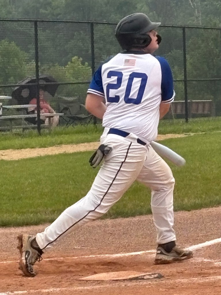 A baseball player wearing a white and blue jersey with the number 20 swings a bat in a grassy field during a game, with blurry spectators and trees in the background.