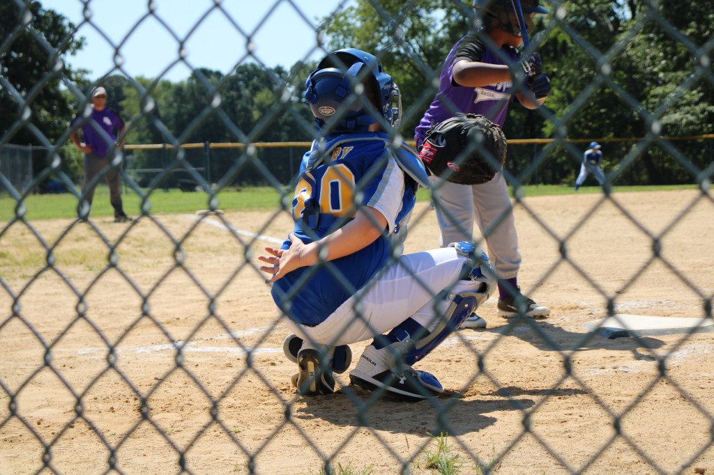 A young baseball player in a blue and white uniform crouches behind home plate, wearing a catcher's mask and gear, while a batter in a black shirt prepares to hit the ball. The scene is framed by a chain-link fence, with other players visible in the background.