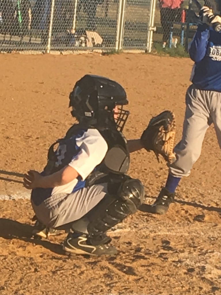 A young baseball catcher in a black helmet and chest protector crouches in a dirt infield preparing for the play, with a player in a blue uniform holding a bat in the background.