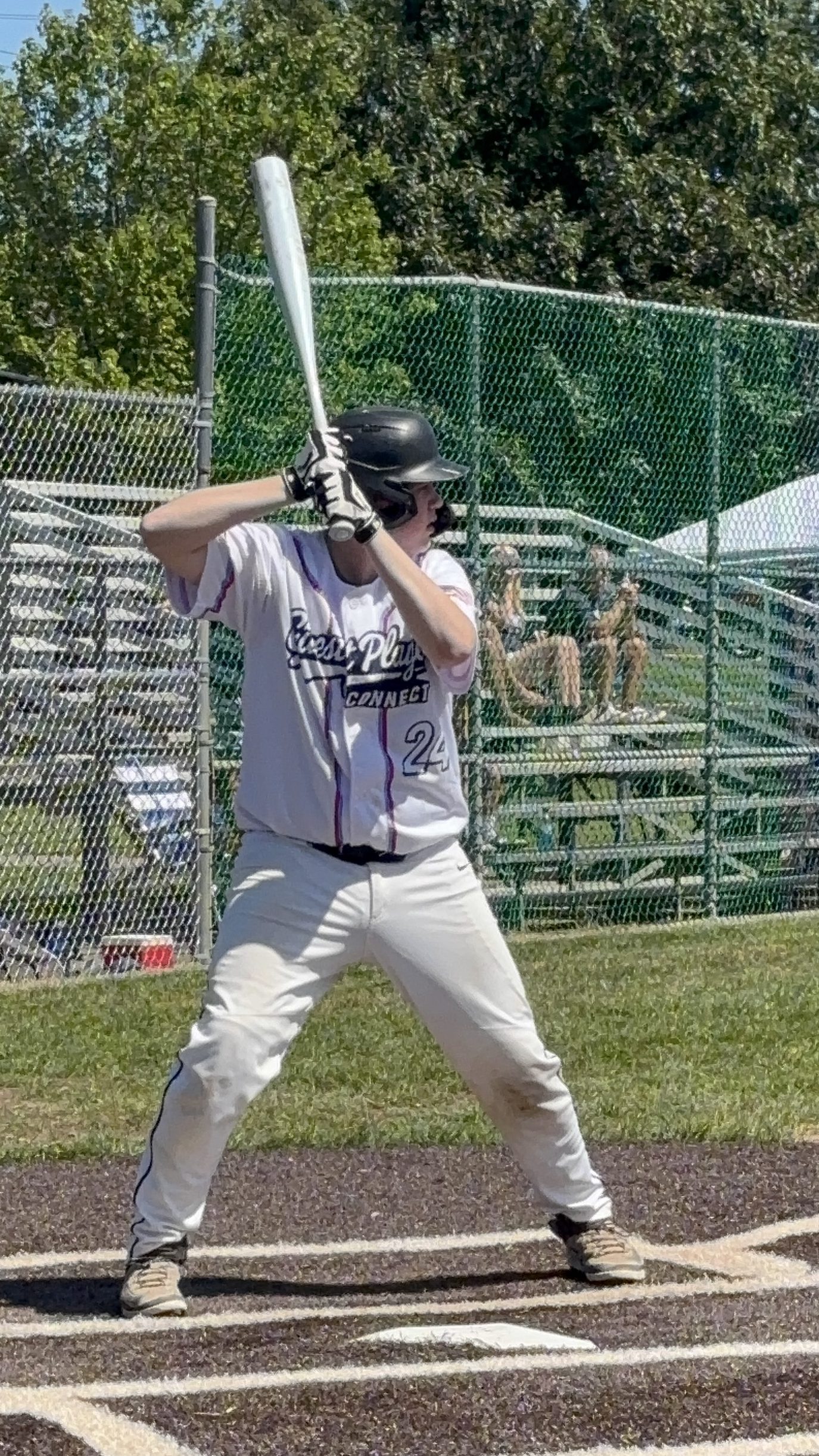 A young baseball player stands at home plate, ready to bat, wearing a white jersey with a black helmet and number 24. In the background, spectators can be seen sitting on bleachers.