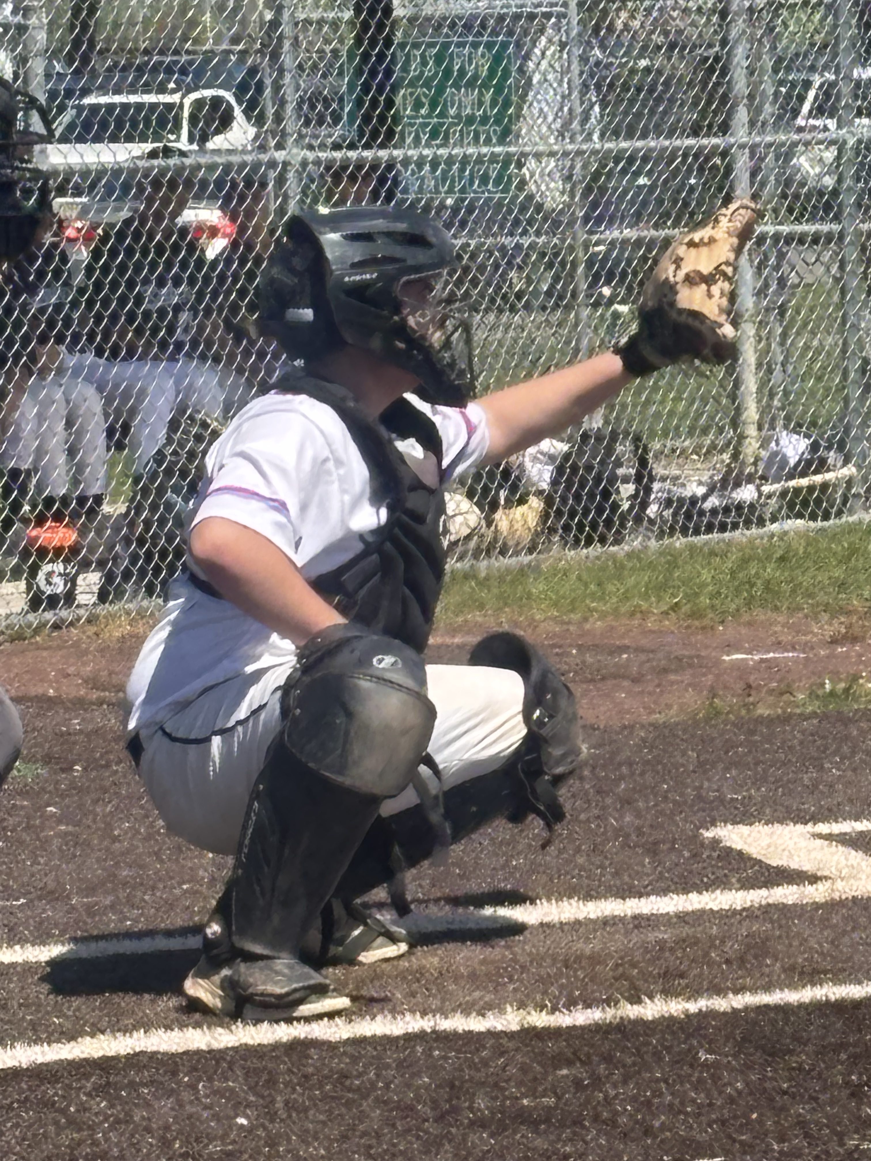 A young baseball catcher in a black helmet and chest protector crouches behind home plate, signaling to the pitcher during a game.