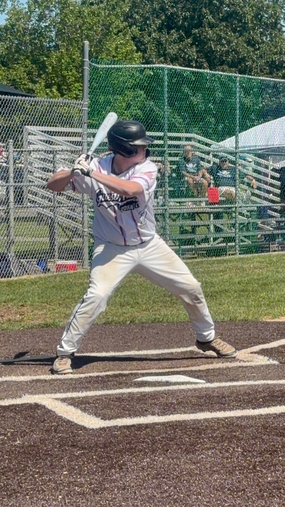 A young baseball player in a white uniform with black accents stands in a batting stance at home plate, holding a bat and preparing to hit. The scene is set on a sunny day with spectators in the background.