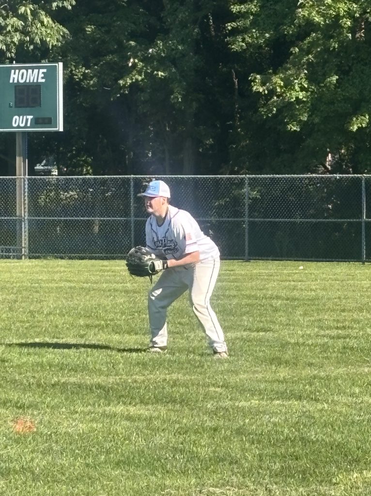 A young baseball player in a gray uniform and blue cap is positioned in the outfield, preparing for a play. A scoreboard is visible in the background showing the home score.