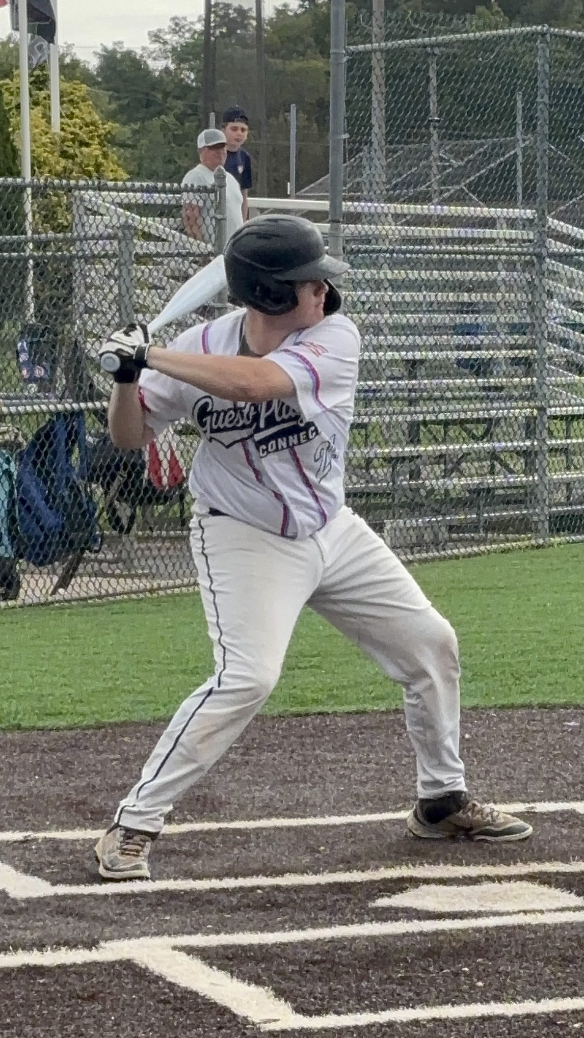 A young baseball player in a white jersey with 'Guest Players' printed on it is in a batting stance at home plate, holding a bat ready to swing. The background features some spectators and bleachers.