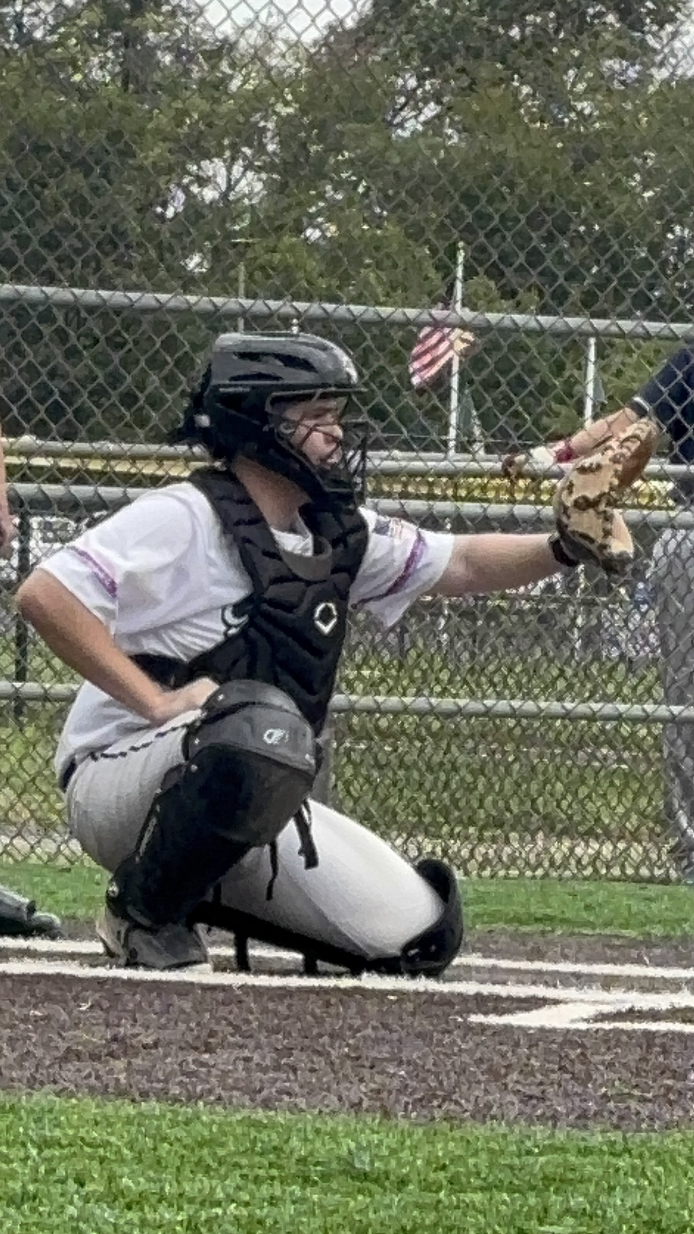 A youth baseball catcher wearing a black helmet and chest protector crouches behind home plate, preparing to receive a pitch. The background shows a baseball field with a chain-link fence and an American flag in the distance.