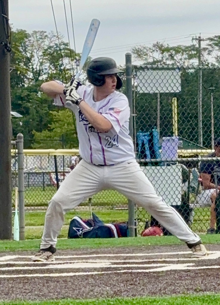 A young baseball player in a batting stance, wearing a black helmet and a white jersey with purple accents. He is positioned in a baseball diamond, ready to hit with a bat.