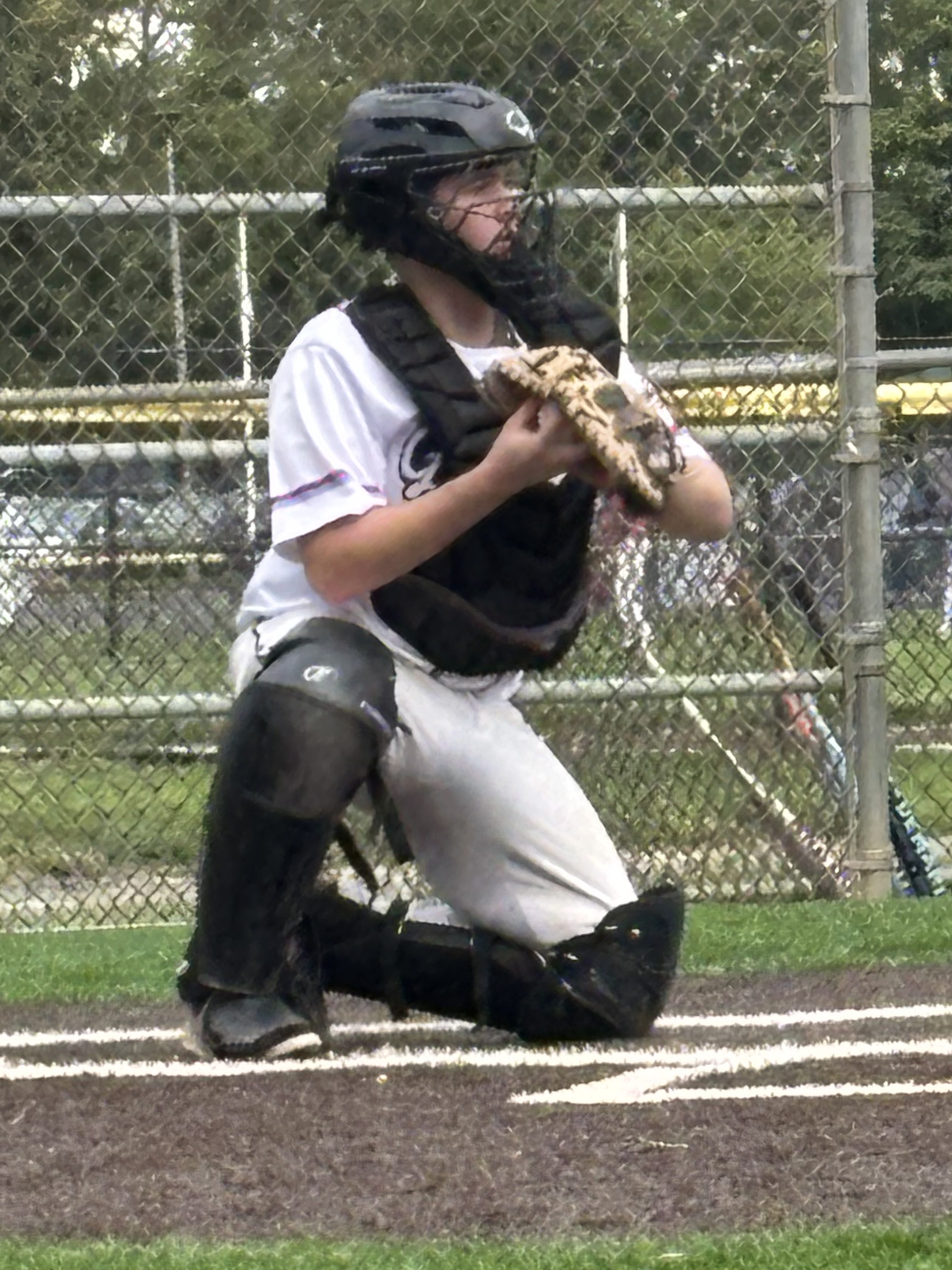 A young baseball catcher in full gear kneeling behind home plate, holding a glove, with a baseball field and fence visible in the background.
