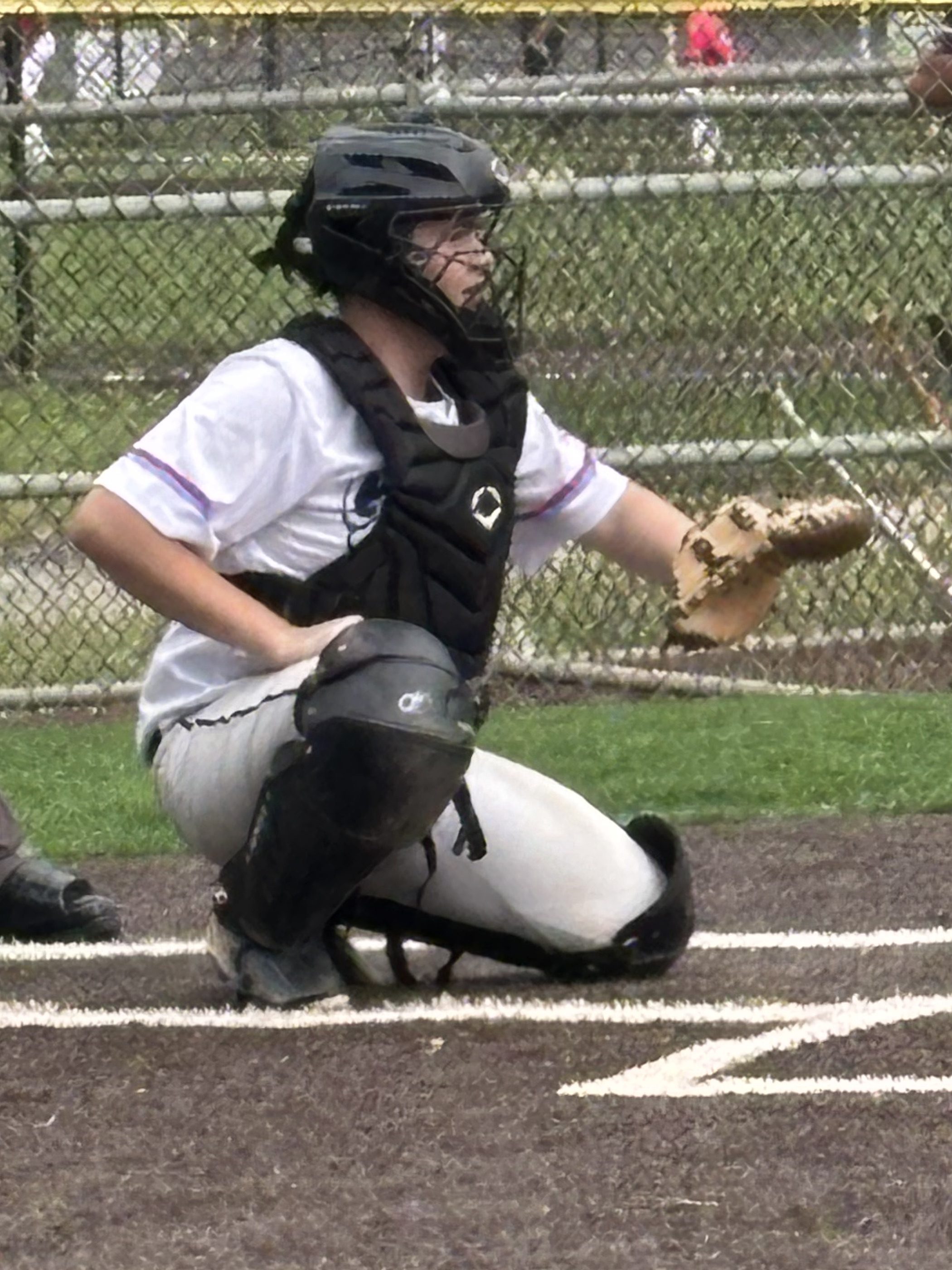 A young baseball catcher in a black helmet and chest protector, crouching behind home plate, with a glove in hand and looking towards the pitcher.
