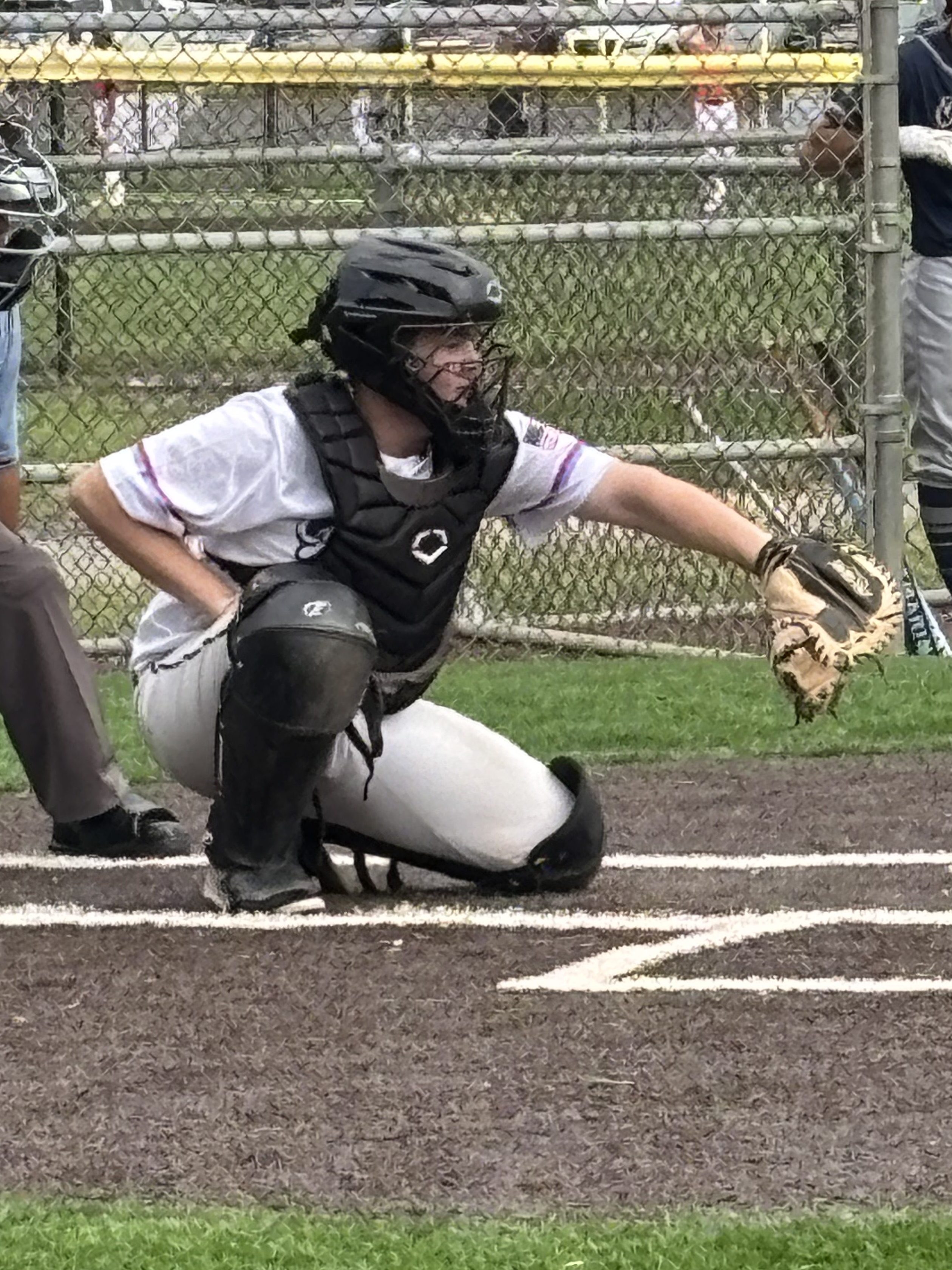 A youth baseball catcher in a black helmet and chest protector crouches behind home plate, ready to receive a pitch.