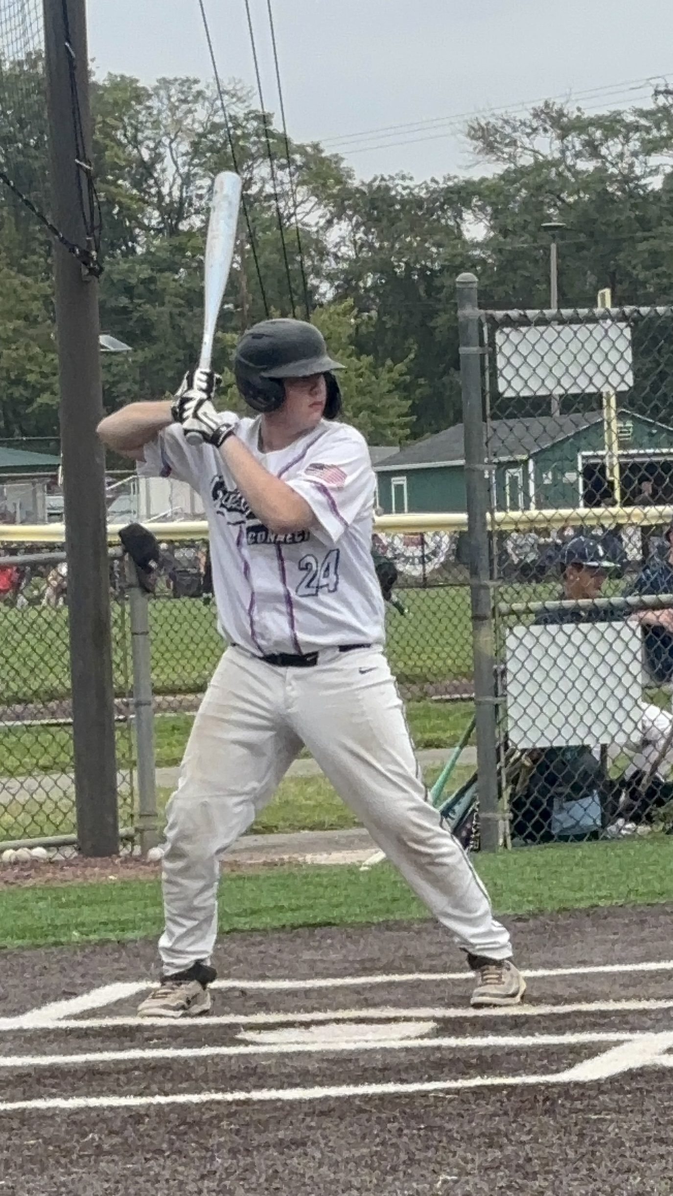 A young male baseball player stands at home plate holding a bat, ready to swing, while wearing a black helmet and a white jersey with the number 24.