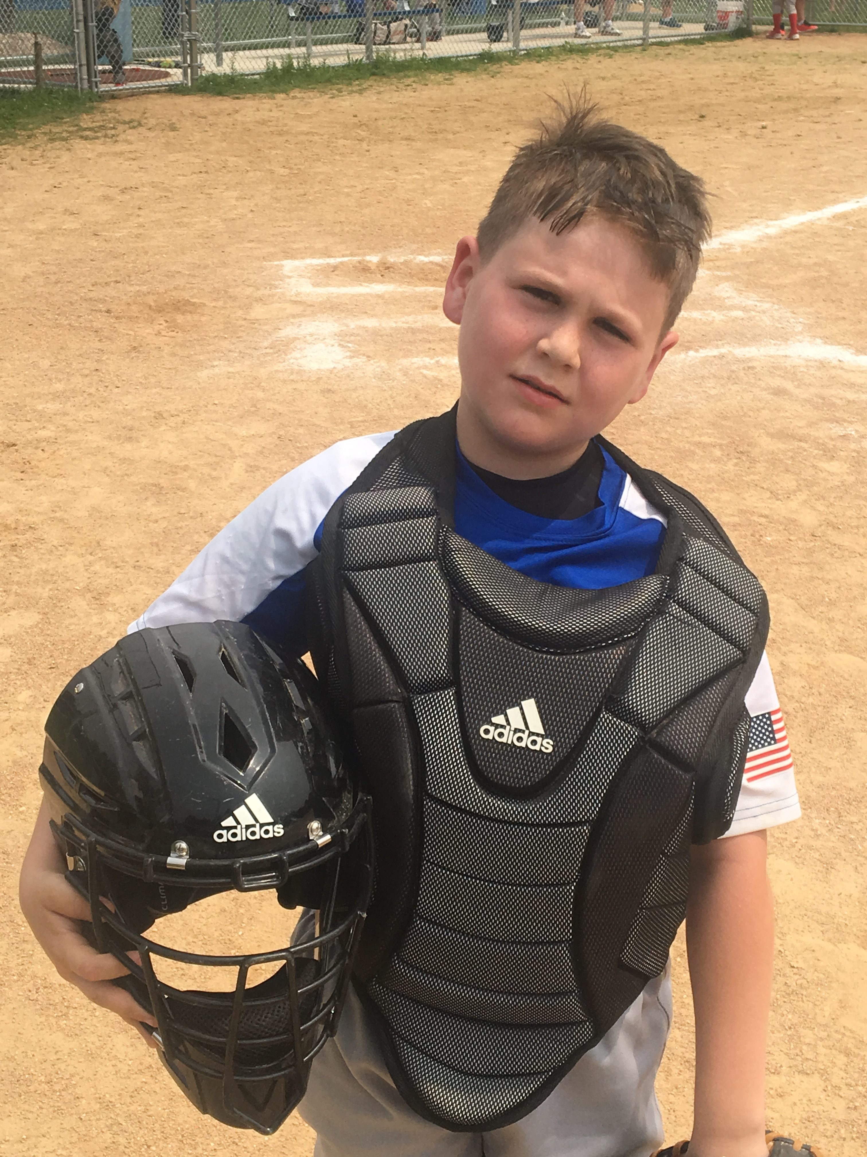 A young baseball catcher wearing a black chest protector and holding a black helmet, standing on a sandy baseball field.