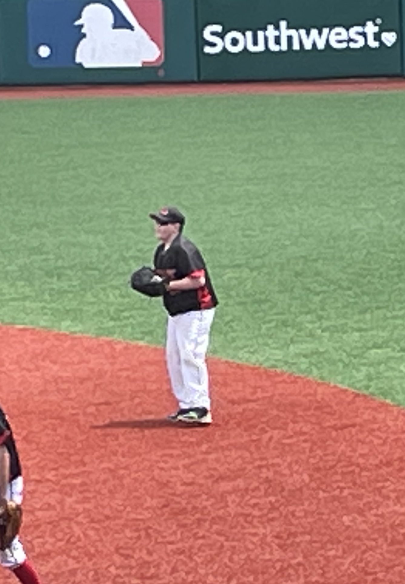 A young baseball player standing on the field, wearing a black jersey with red accents and white pants, holding a glove and preparing to play.