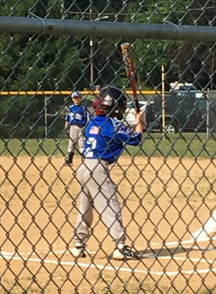 A young boy in a blue baseball uniform prepares to bat on a baseball field with a chain-link fence in the foreground. Another boy stands in the background.