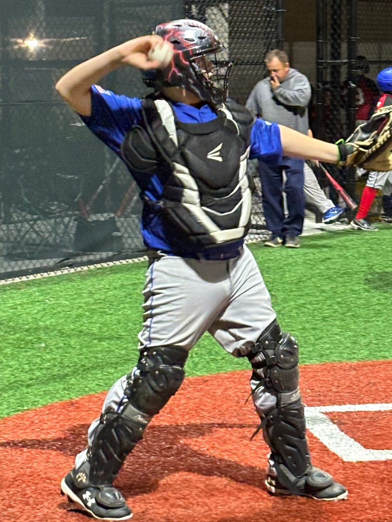 A young baseball catcher in a black and white catcher’s gear throws a baseball, wearing a blue jersey and standing on a baseball field with artificial turf.