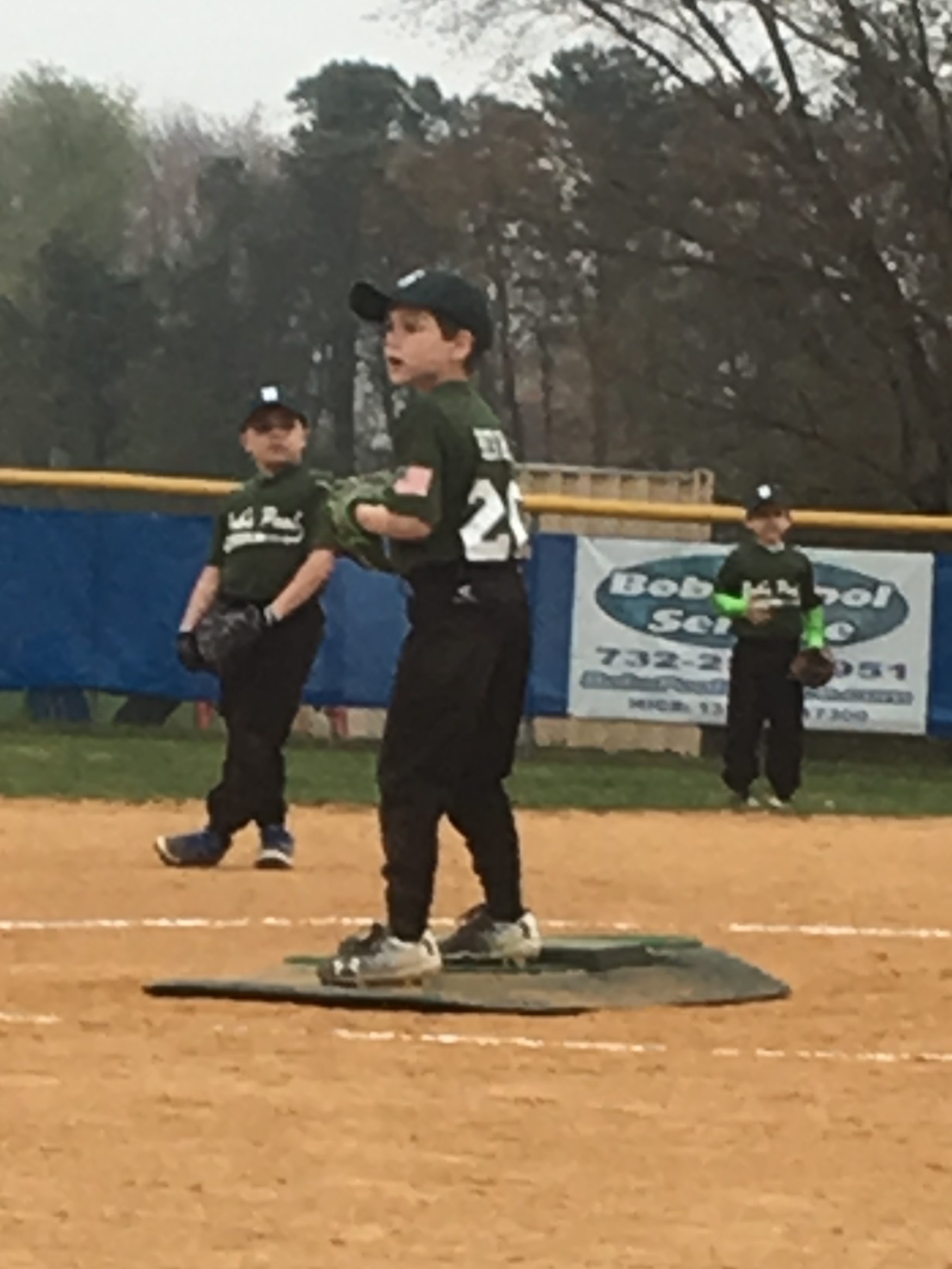A young baseball player on the pitcher's mound wearing a green jersey and cap, preparing to throw, with teammates in the background.