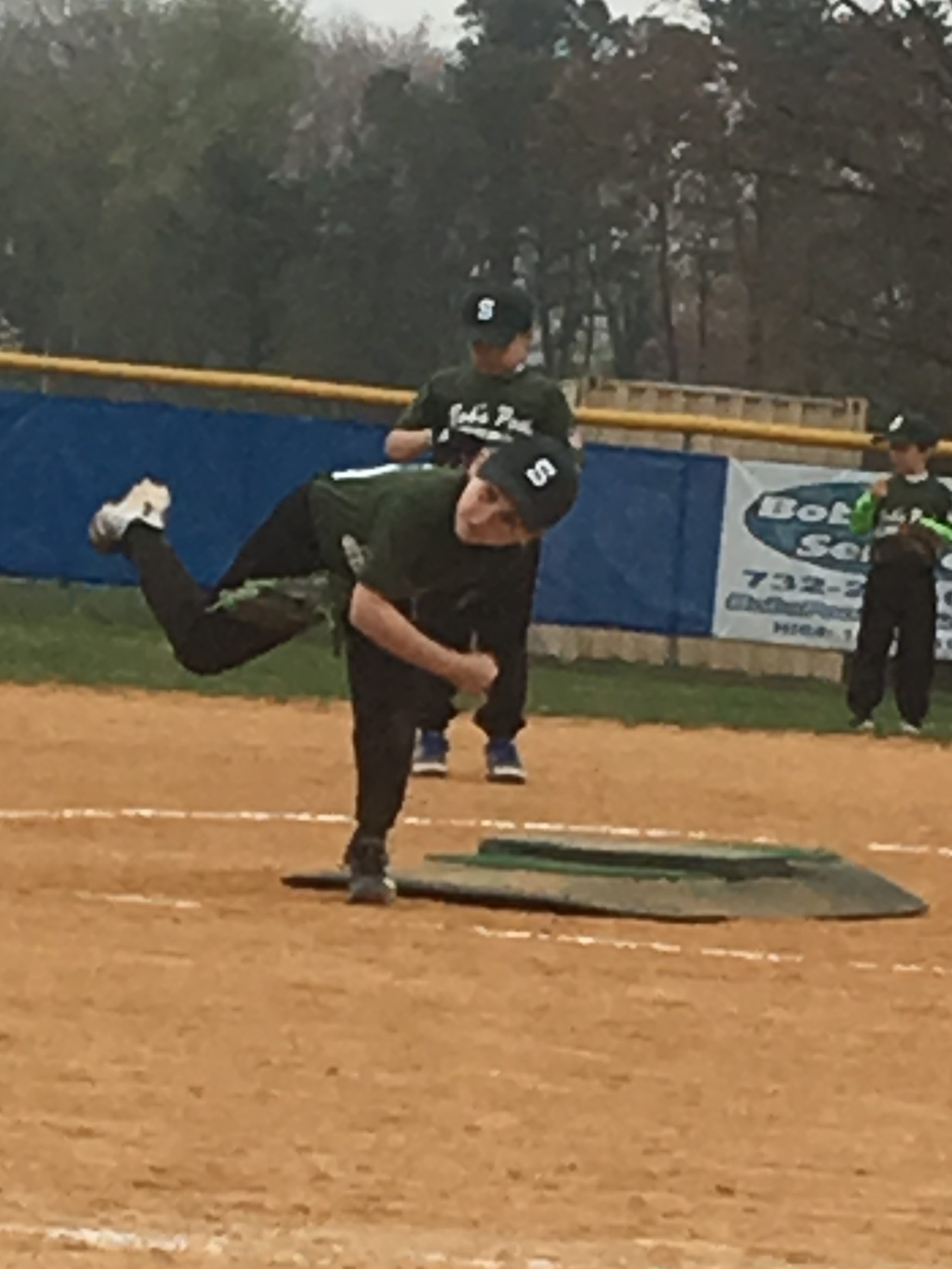 A young baseball player in a green jersey pitches from the mound on a sandy field, with teammates in the background.