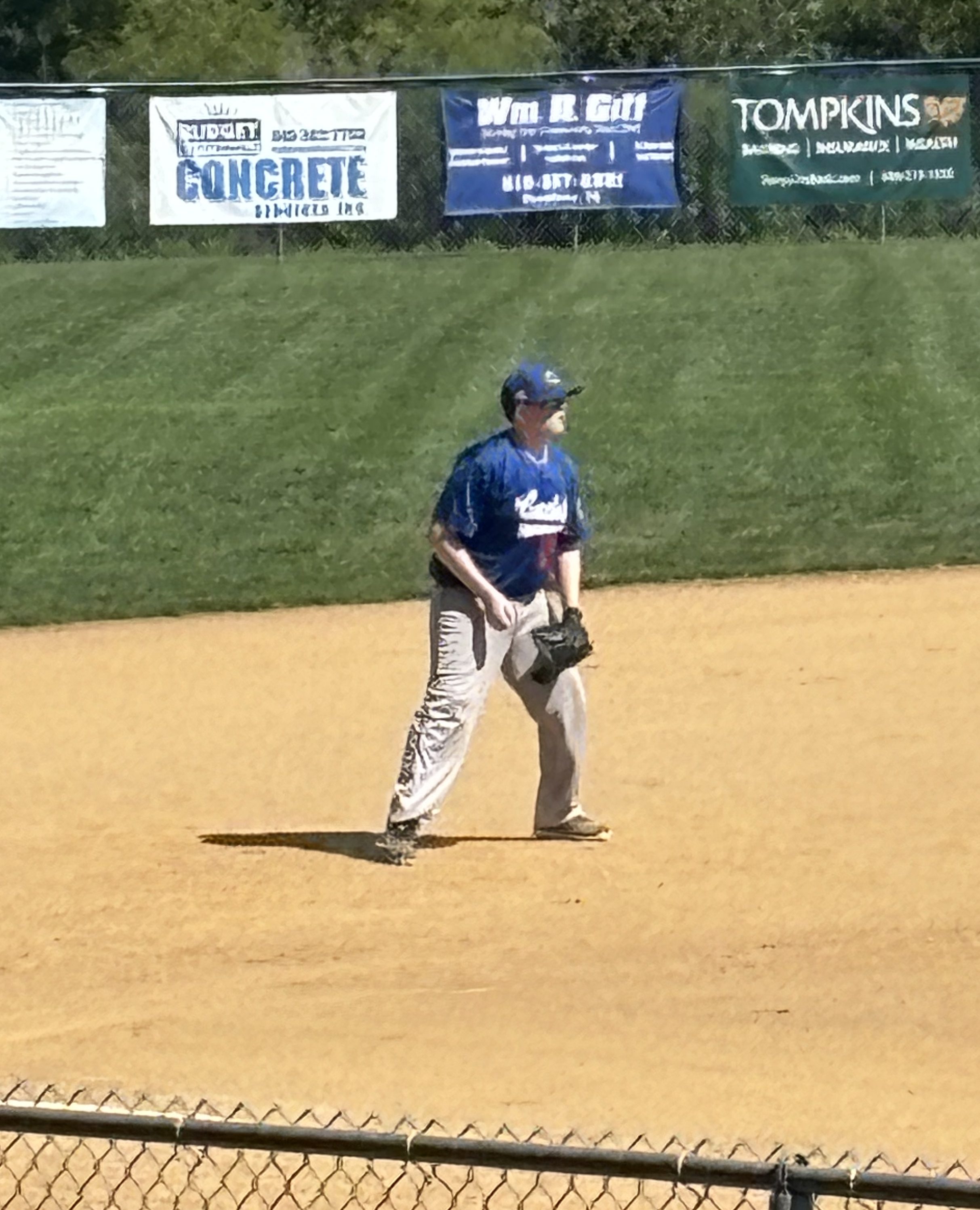 A baseball player in a blue jersey and gray pants stands on a sandy infield, preparing to play. Advertising banners are visible in the background, along with a grassy area.