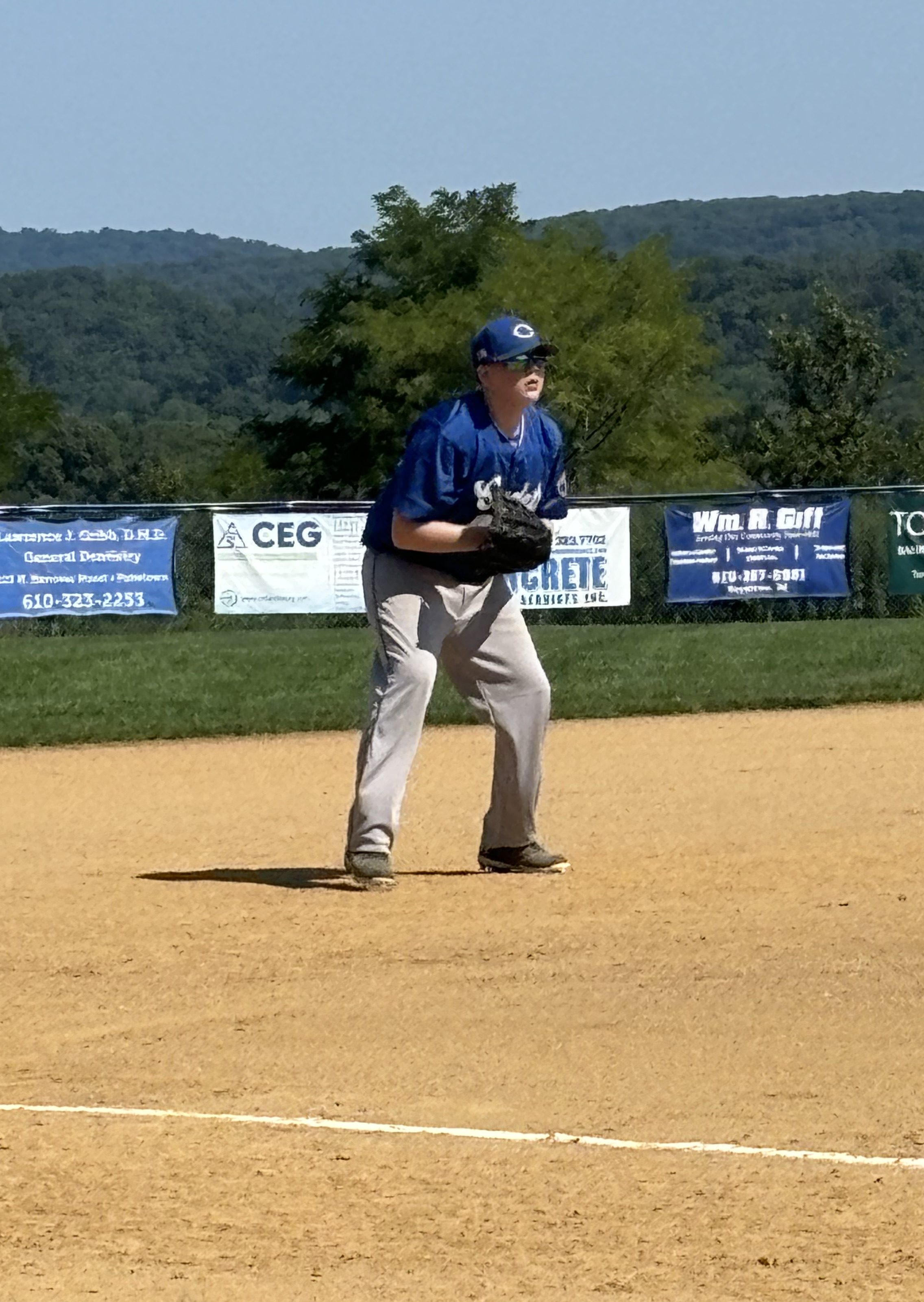 A young baseball player in a blue jersey and grey pants is in a pitcher's stance on a sandy baseball field. He is preparing to make a play, with mountains visible in the background and sponsor banners along the outfield fence.