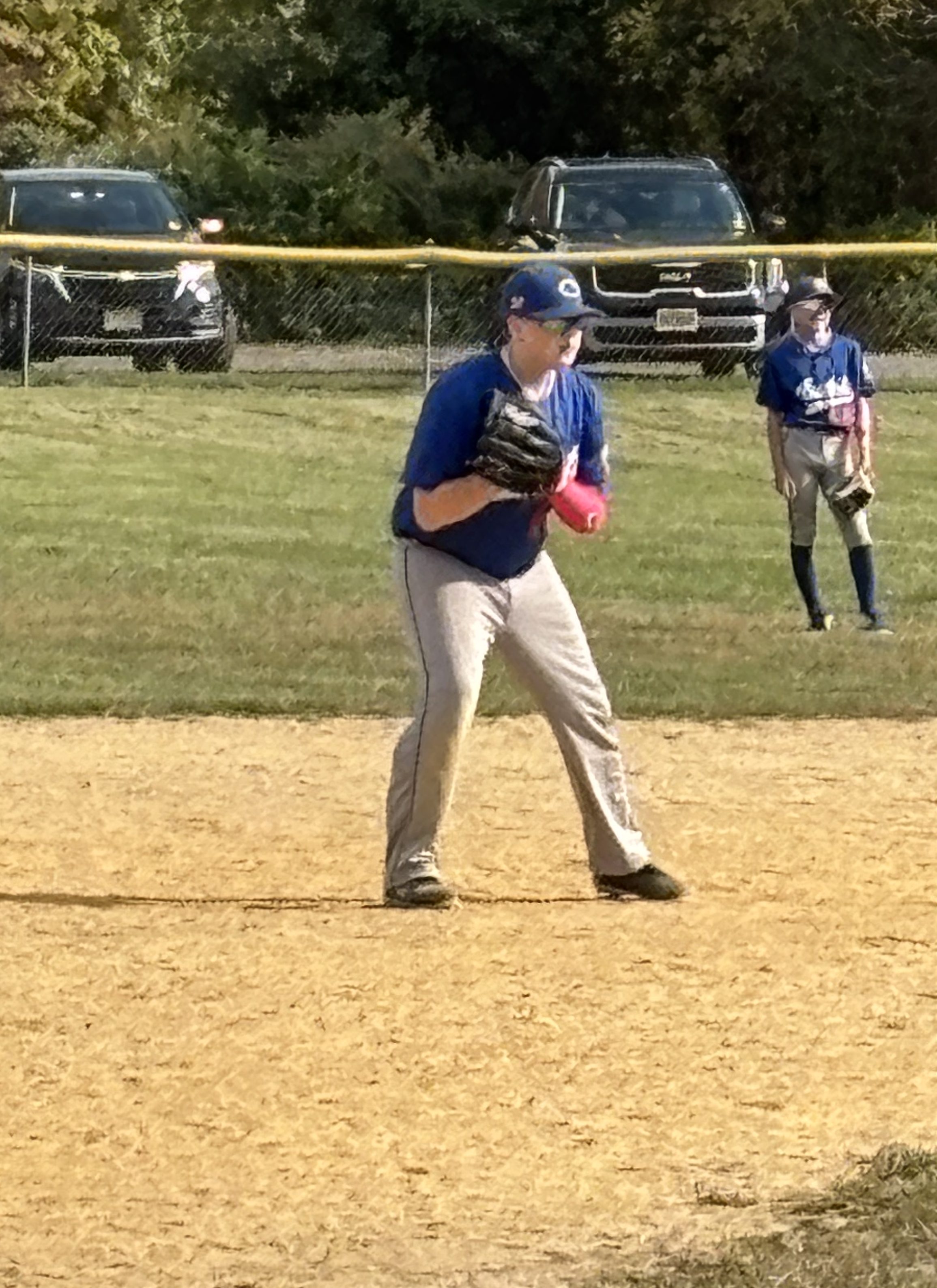 A young baseball player in a blue shirt and gray pants stands ready on the field, wearing a glove on his left hand and a pink sleeve on his right arm. In the background, two cars are parked near a fence, and another player in a blue shirt stands off to the side.