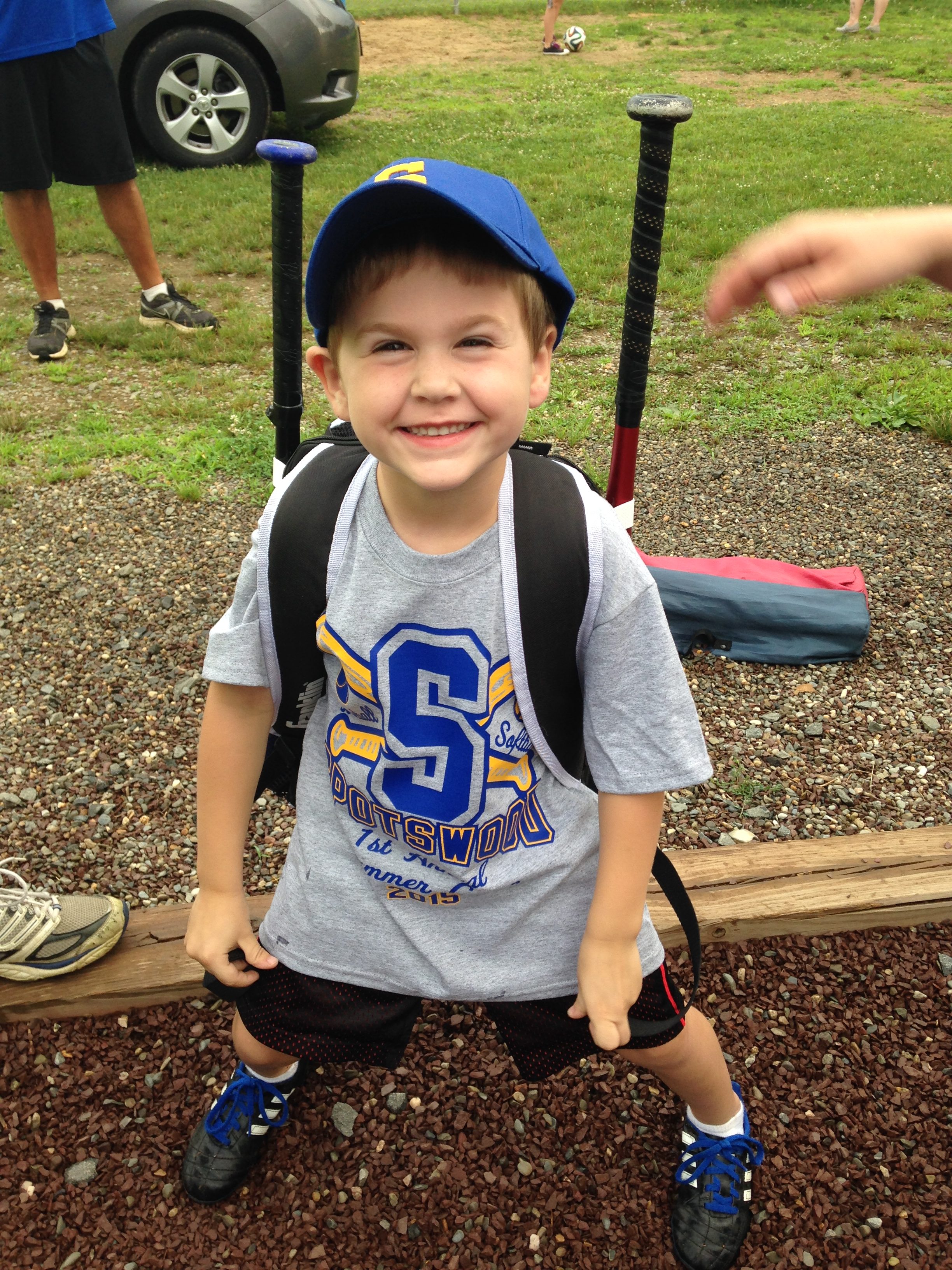 A smiling young boy wearing a blue cap and a gray t-shirt with a sports logo, standing on gravel ground, with a backpack and baseball gear visible in the background.