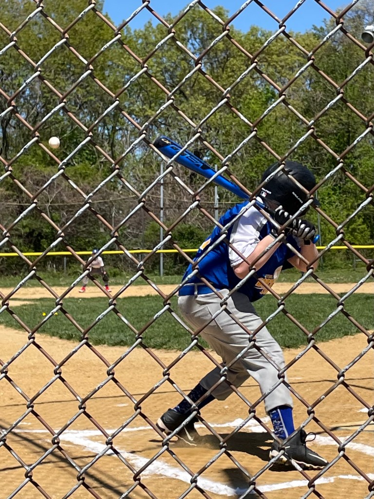 A young baseball player in a blue jersey prepares to hit a ball, viewed through a chain-link fence on a sunny day at the baseball field.