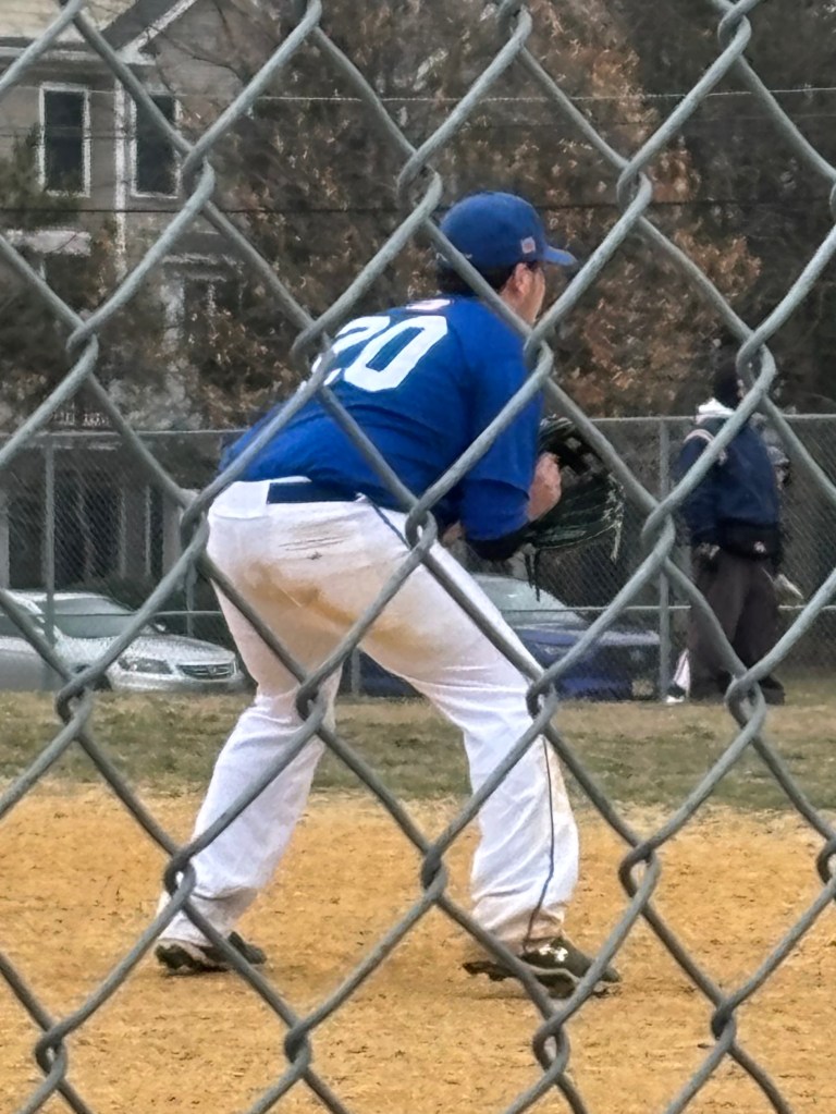 A baseball player in a blue jersey and white pants is crouching in a ready position on a baseball field, with a chain-link fence in the foreground.