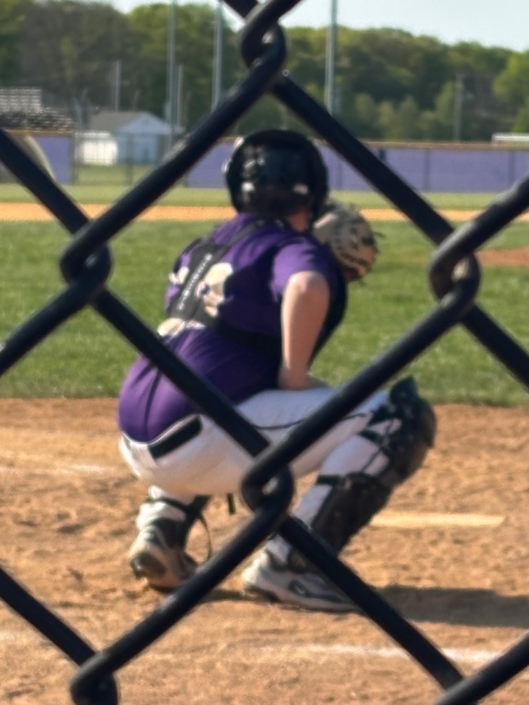A youth baseball player in a purple jersey and white pants squatting behind home plate, wearing a catcher's helmet and gear, with a focus on the player's back through a chain-link fence.