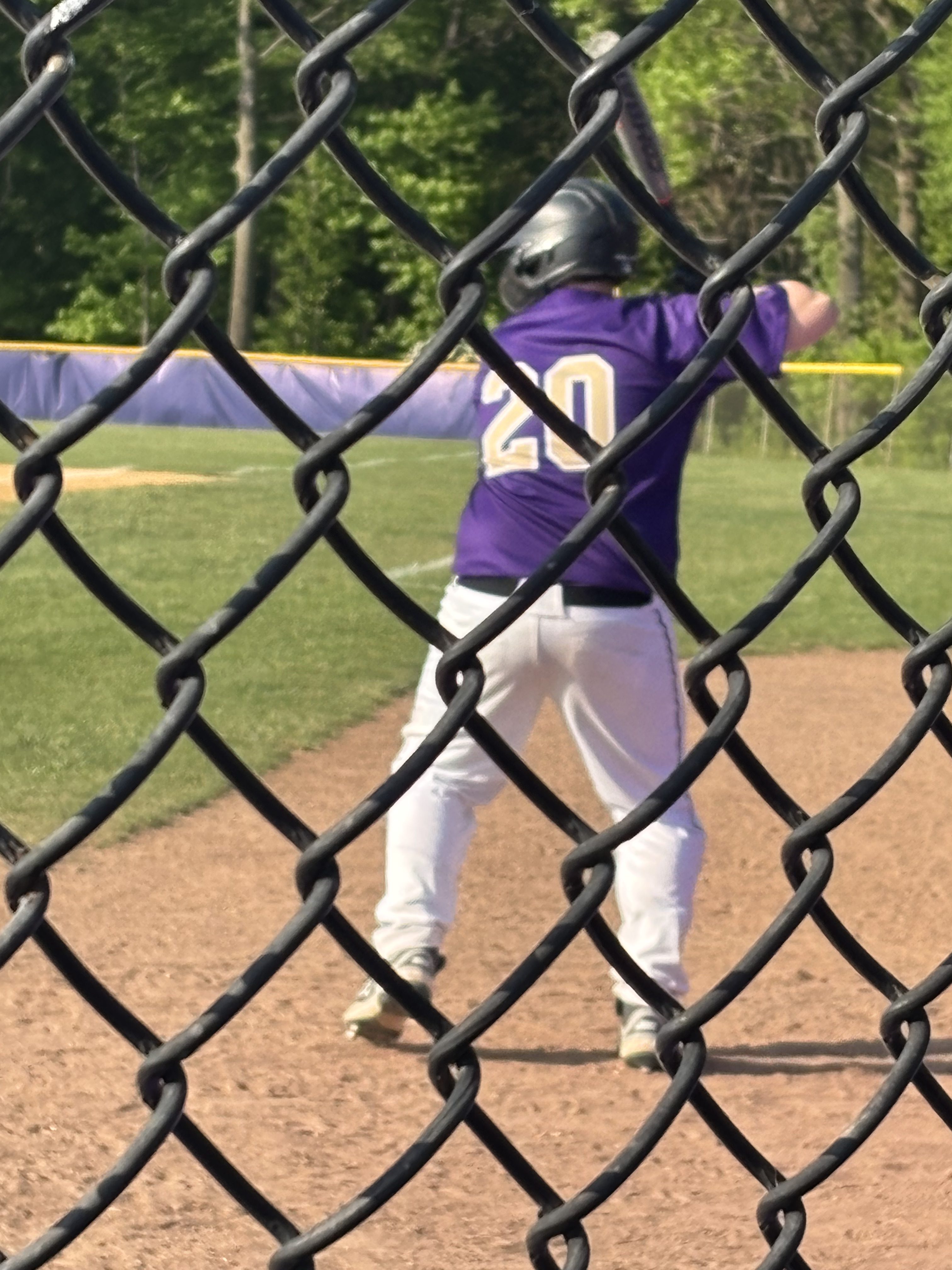 A baseball player in a purple jersey with the number 20 stands at the plate, preparing to bat, with a chain-link fence in the foreground.
