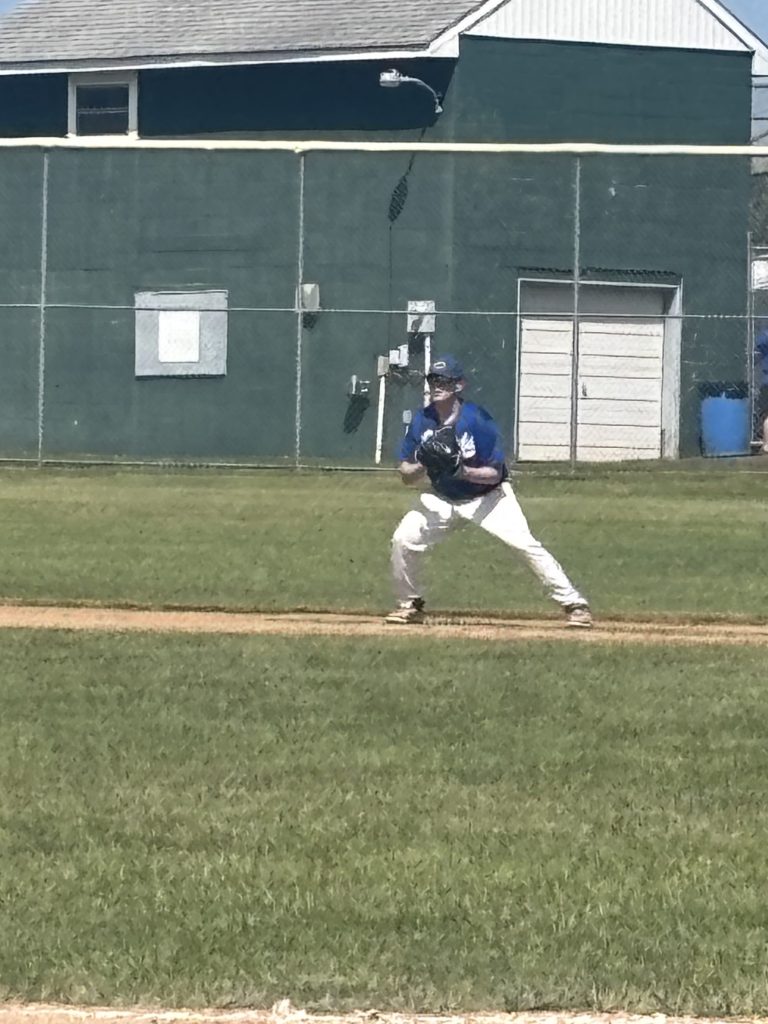 A baseball player in a blue jersey and white pants is positioned defensively on the field, ready to catch a ball, with a green building and grass in the background.