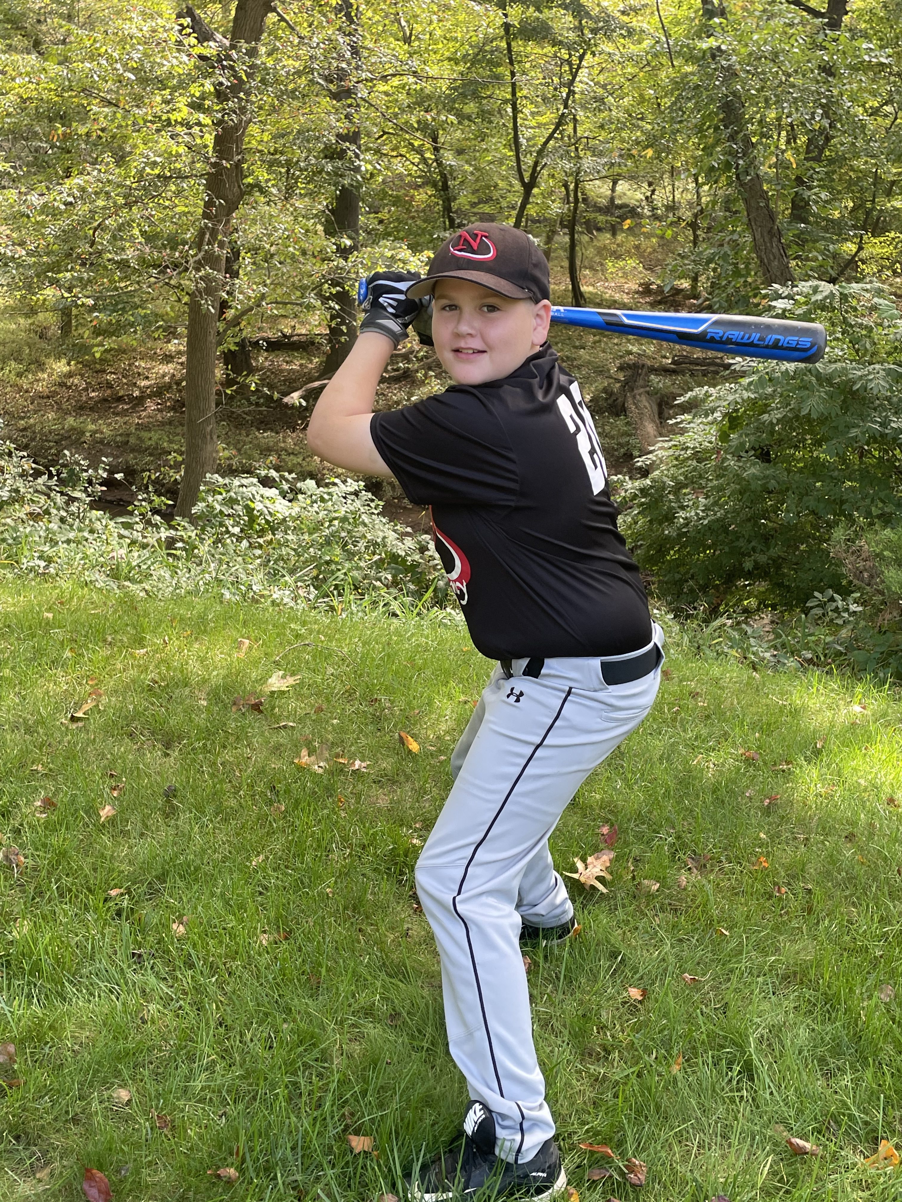 A young boy in a black baseball uniform with the number 23 stands in a grassy area, holding a blue baseball bat and preparing for a swing. Trees and foliage are visible in the background.