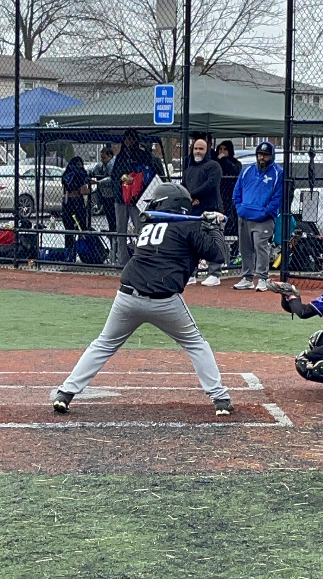 A young baseball player in a black jersey and gray pants stands in the batter's box, preparing to bat with a baseball bat. The background shows other players and spectators at a baseball field.