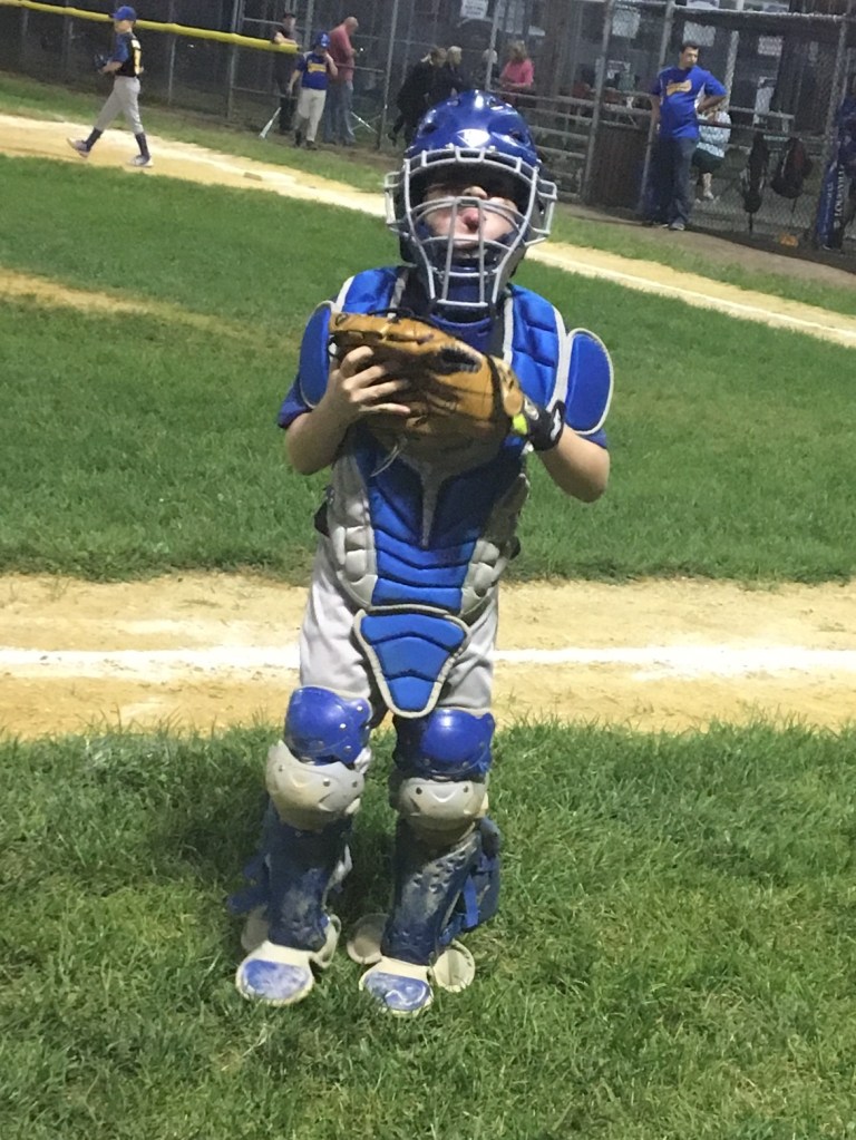 A young baseball player in full catcher gear, standing on a field. The child is wearing a blue helmet and chest protector, holding a baseball glove. In the background, other players and spectators can be seen.