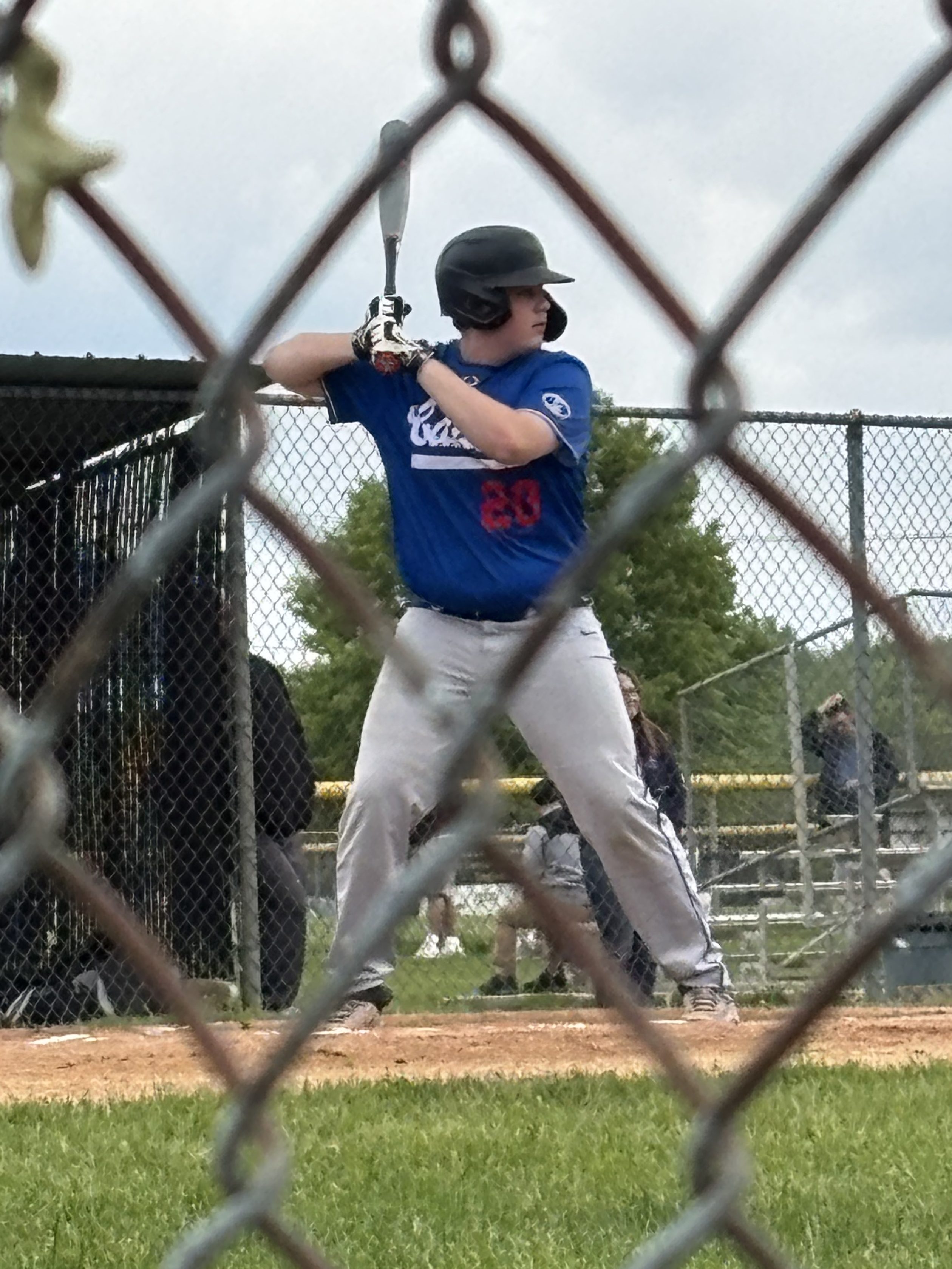 A young baseball player in a blue jersey and black helmet, preparing to bat, with a chain-link fence in the foreground.